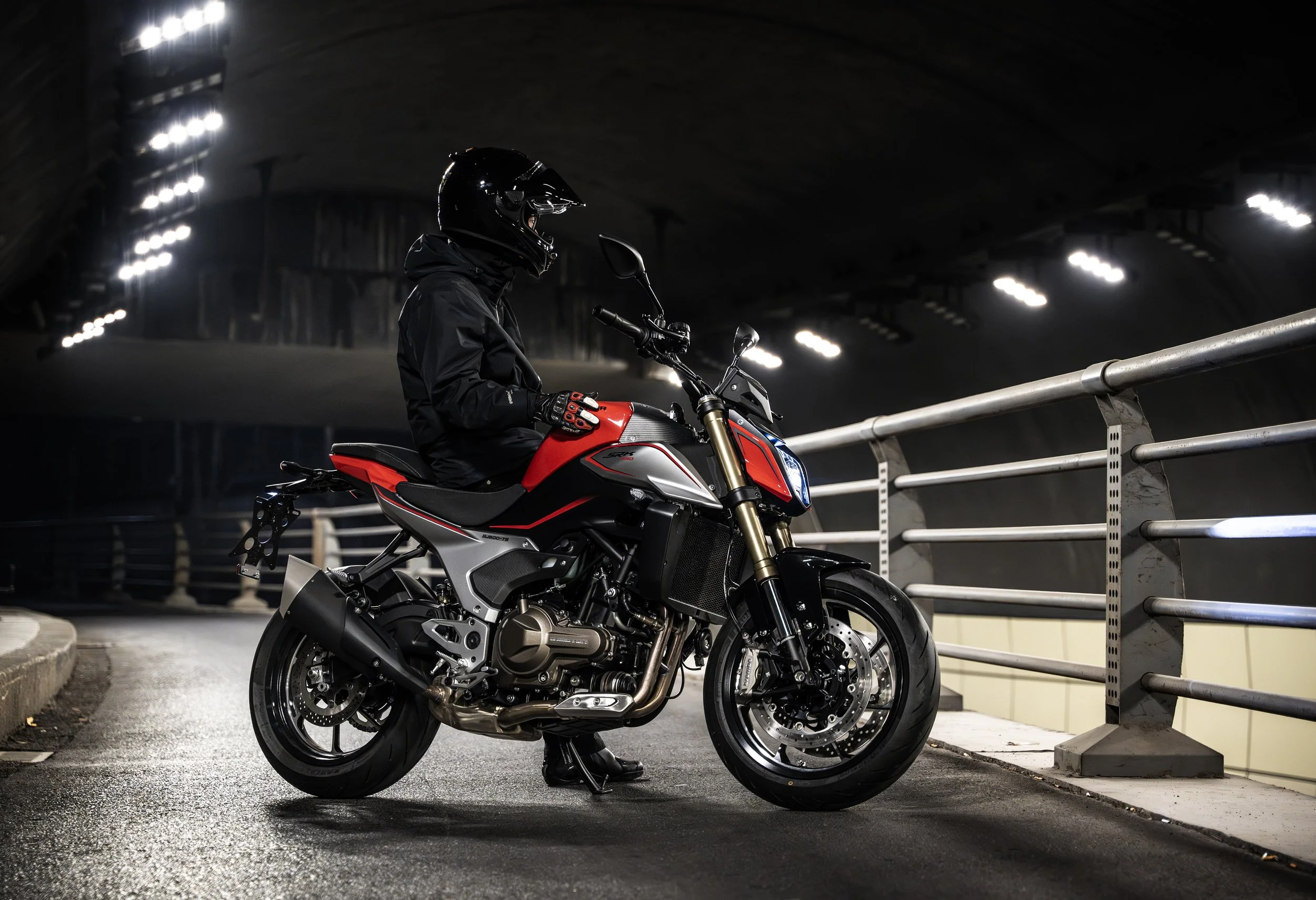 Motard en casque et équipement de protection, assis sur une moto dans un tunnel sombre, éclairé par des lumières.