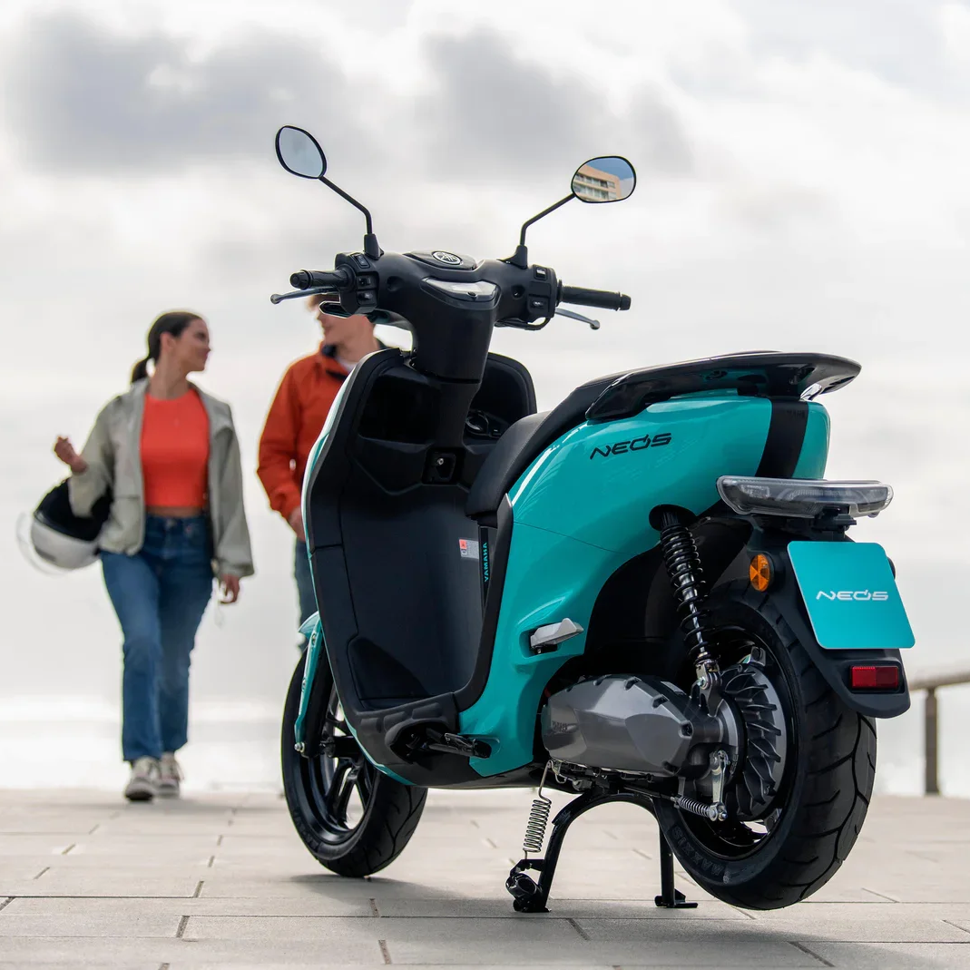 Une moto électrique de couleur turquoise avec le mot 'NEOOS' écrit dessus, stationnée sur un trottoir, avec deux personnes discutant en arrière-plan sous un ciel nuageux.
