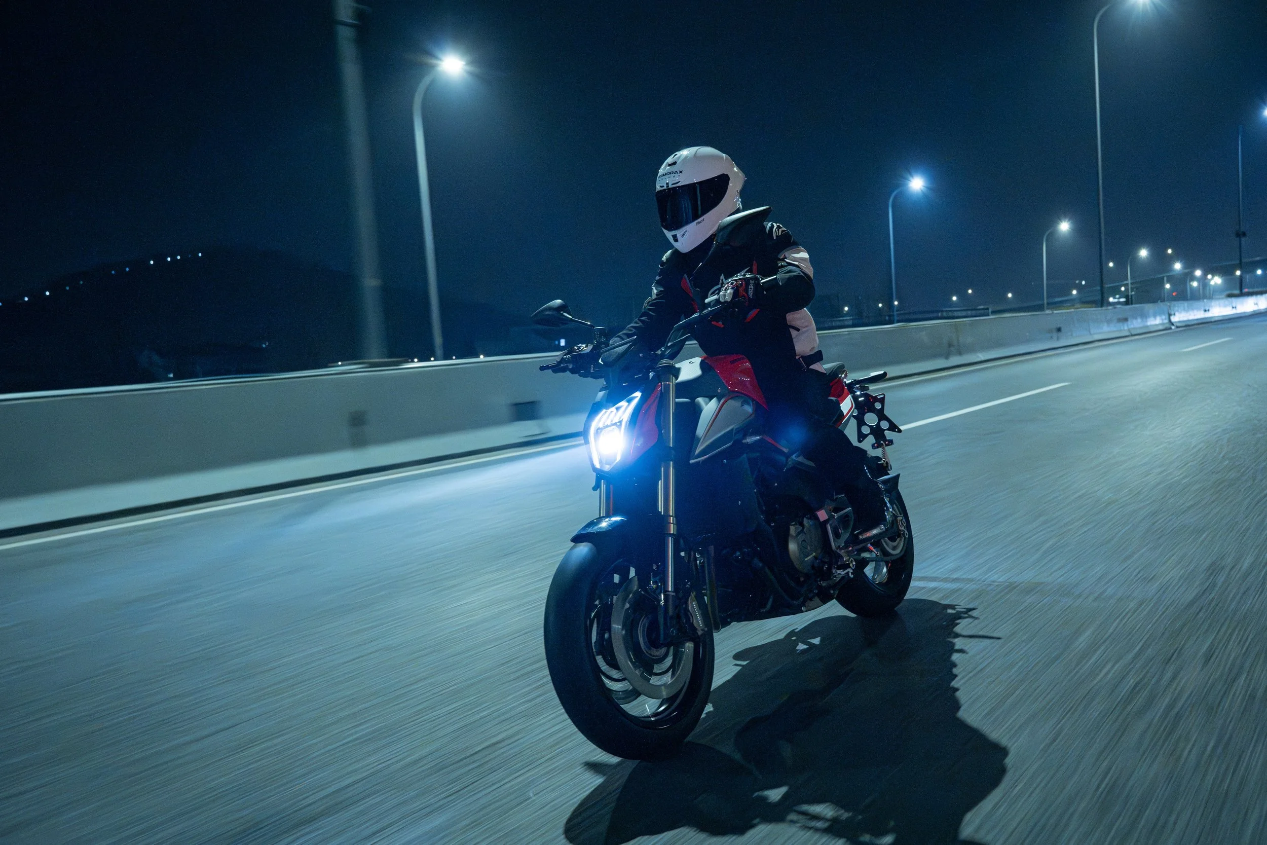 Un motard portant un casque intégral noir et une veste de moto, roulant de nuit sur une route éclairée par des lampadaires, sous un ciel sombre.