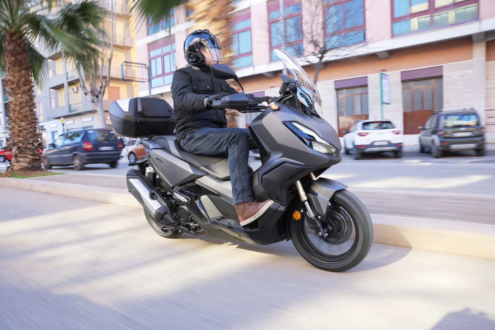 Personne portant un casque à moto roulant à vélo sur une avenue urbaine avec des voitures stationnées et des bâtiments en arrière-plan.