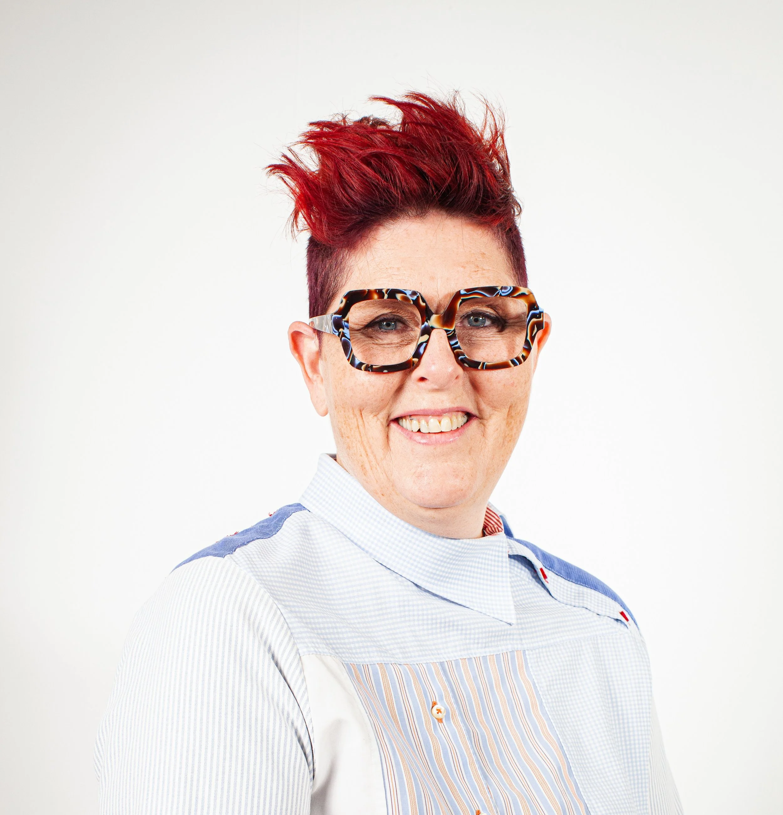 Person with short red hair, wearing large tortoiseshell glasses, smiling, dressed in a light-colored shirt with blue and red accents, against a plain white background.