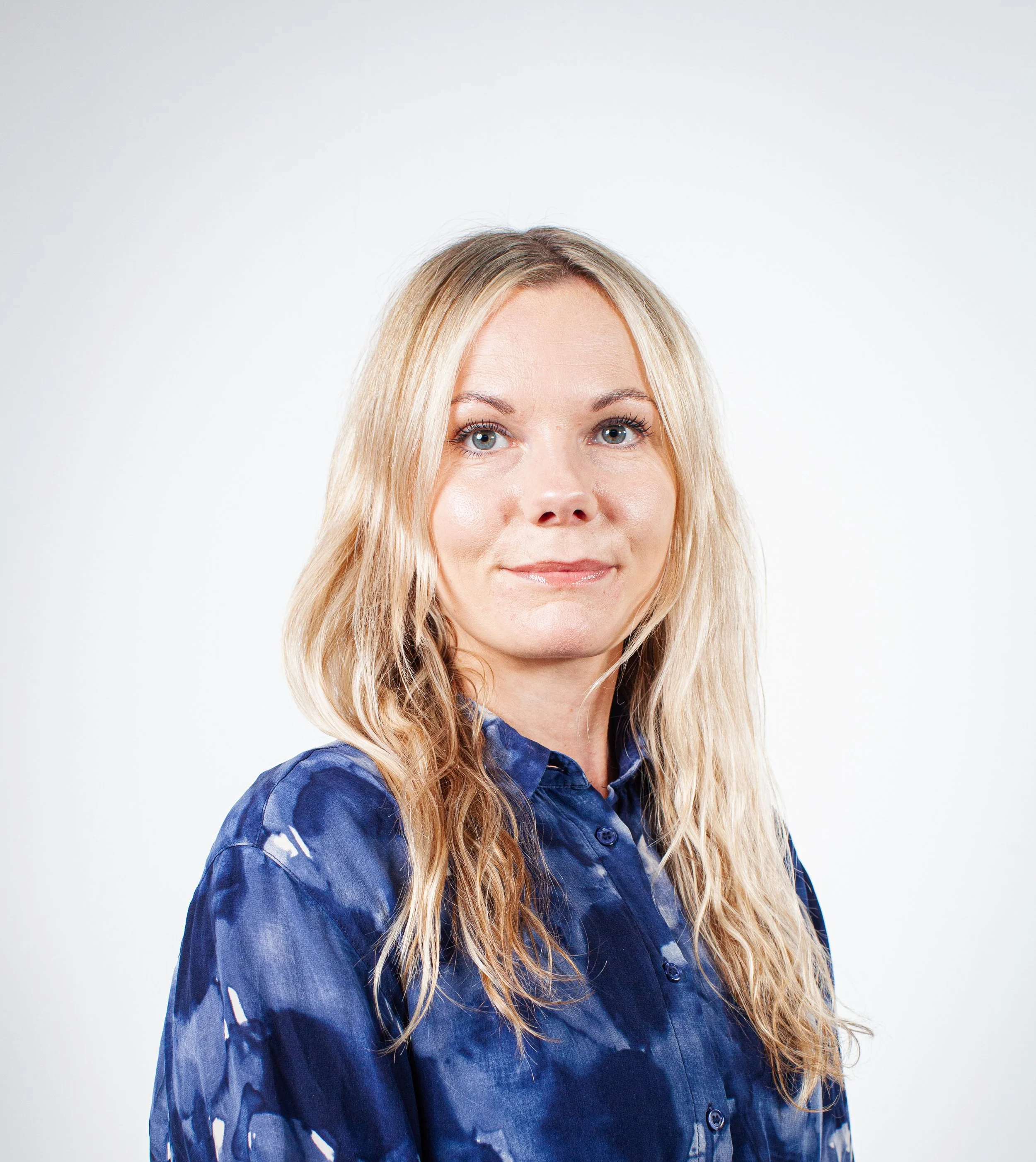 A woman with long blonde hair wearing a blue patterned shirt, looking at the camera with a slight smile.