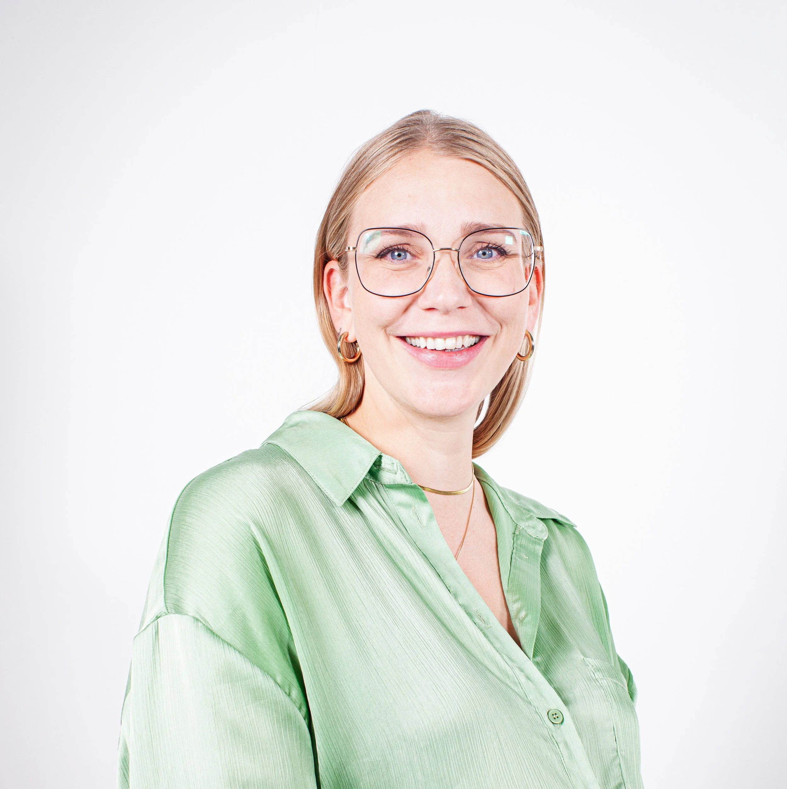 Portrait of a smiling woman with blonde hair, wearing glasses, gold earrings, a gold necklace, and a light green satin blouse against a plain white background.