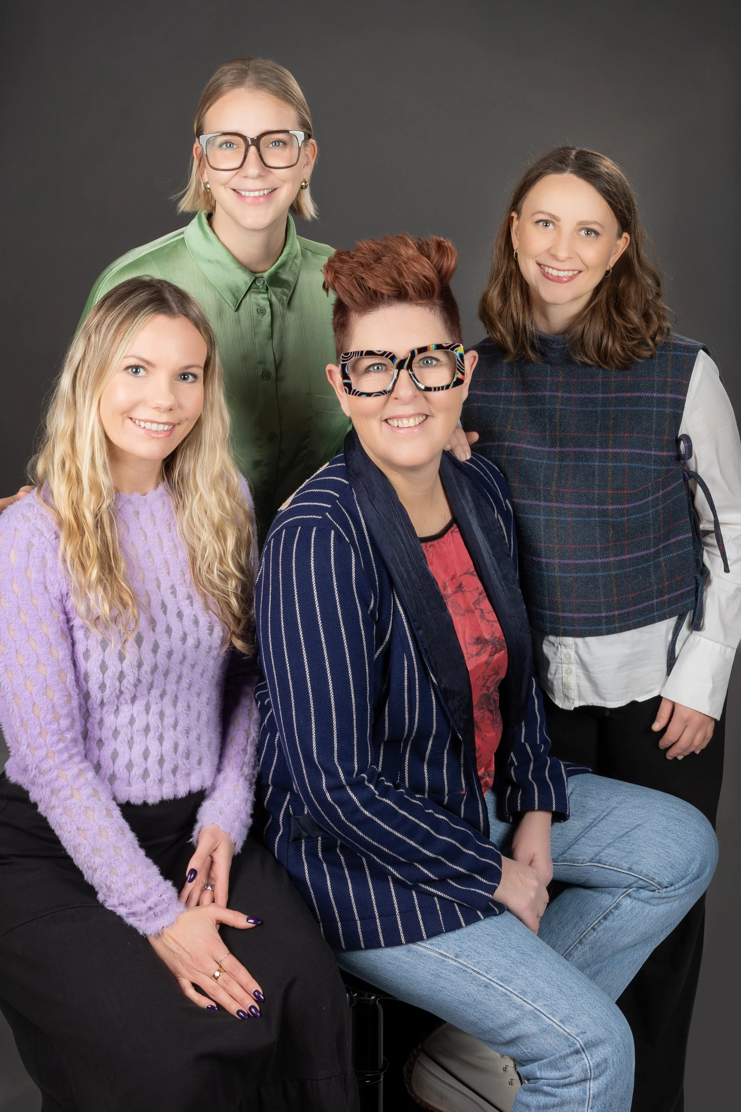 Group portrait of four women smiling, posed against a dark gray background. One woman has short red hair and wears glasses with a zebra print frame, sitting on a stool. Others are standing or sitting nearby, wearing colorful sweaters and shirts.