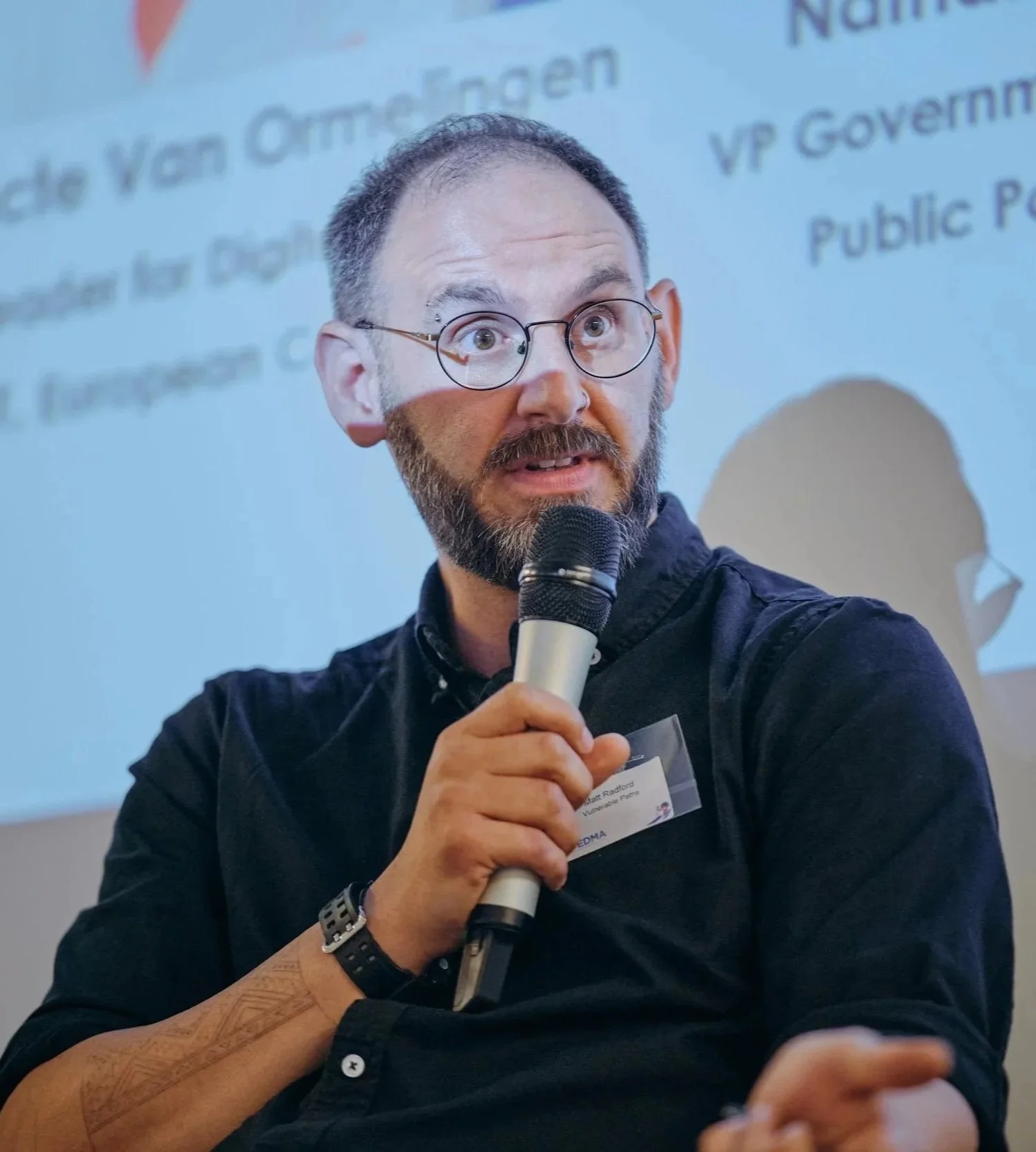 A man with glasses and a beard holding a microphone, speaking at an event. He is wearing a black shirt, a watch, and has a name tag. The background shows a presentation slide with partly visible text.
