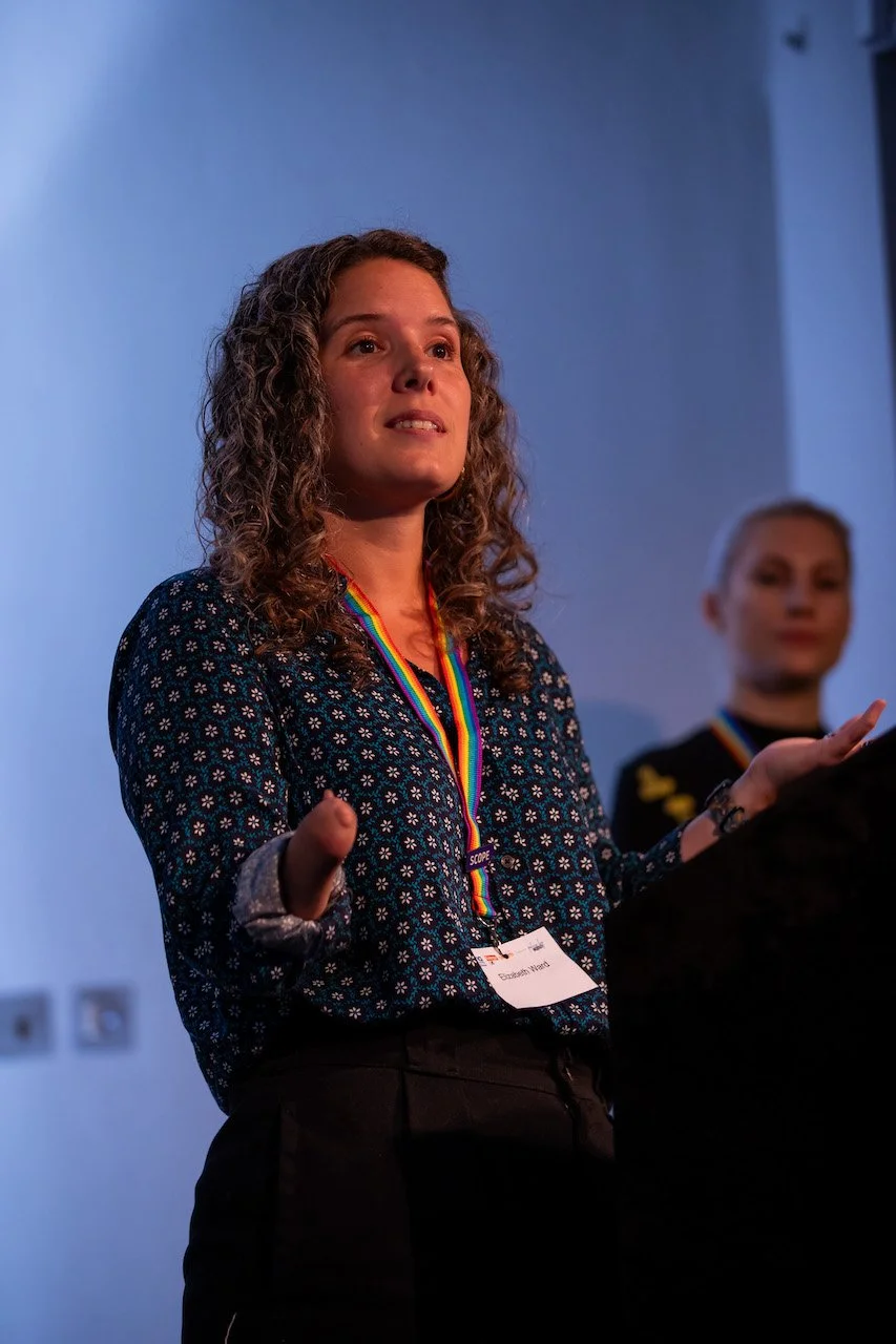 A woman with curly hair speaking at an event, wearing a rainbow medal and a name badge, with another woman blurred in the background.