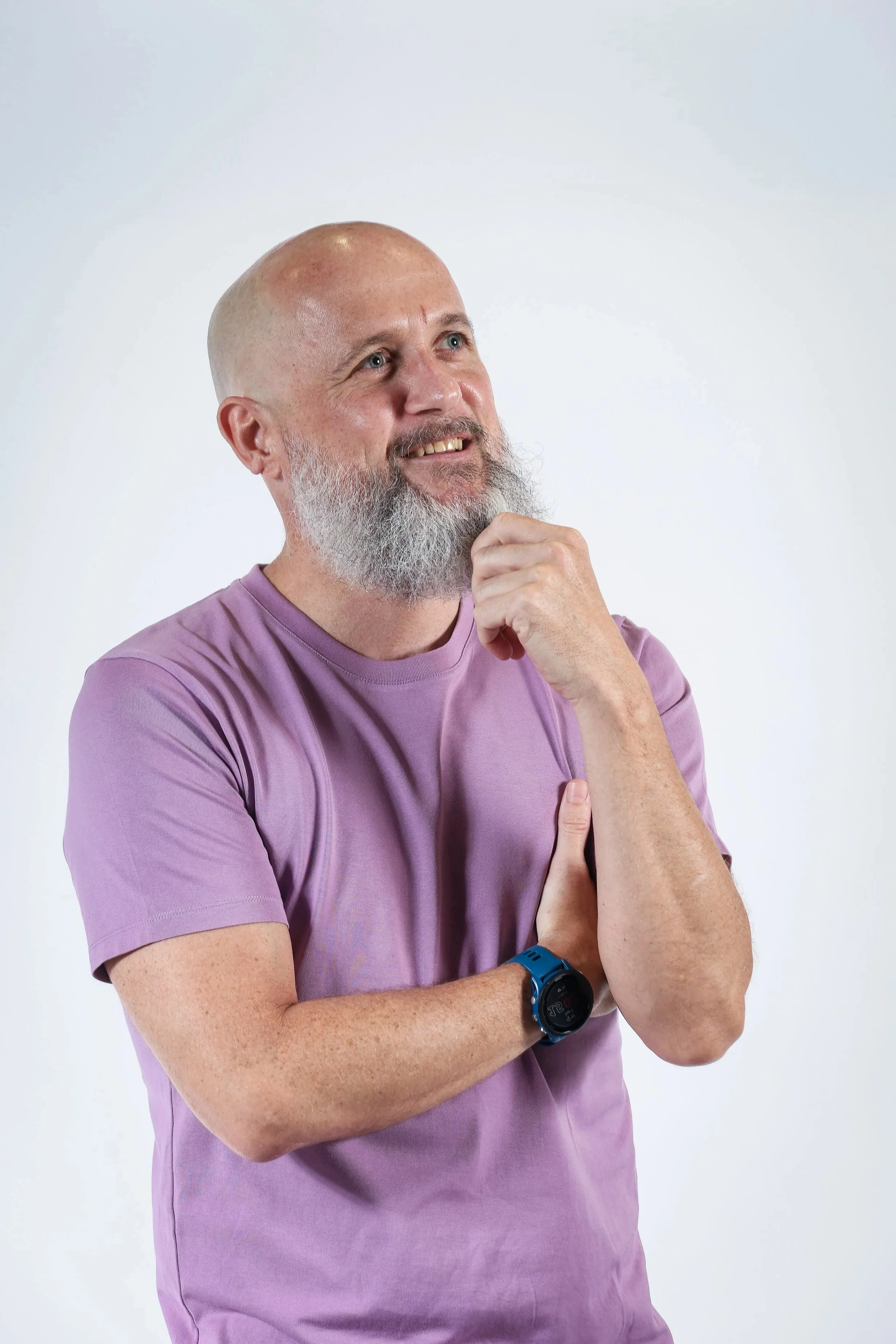 A man with a bald head and a gray beard is wearing a purple t-shirt and a black and blue smartwatch. He is posing with his right hand under his chin and his left arm crossed, looking slightly upward with a thoughtful expression, against a plain white background.