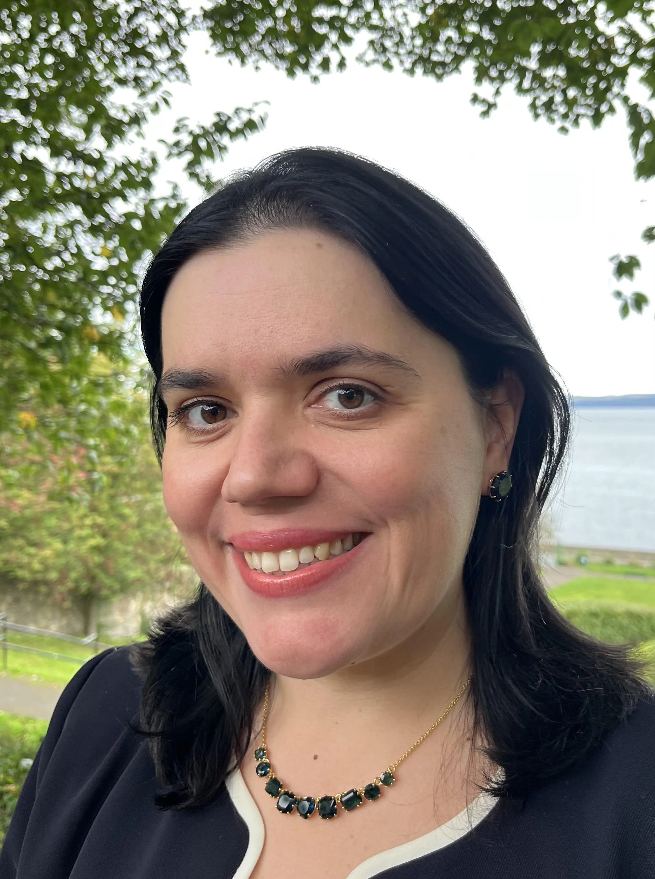 Olga is smiling with black shoulder length hair and light skin tone. She is wearing a purple top, and gold necklace with stones. Behind her is a lake, trees and grass.