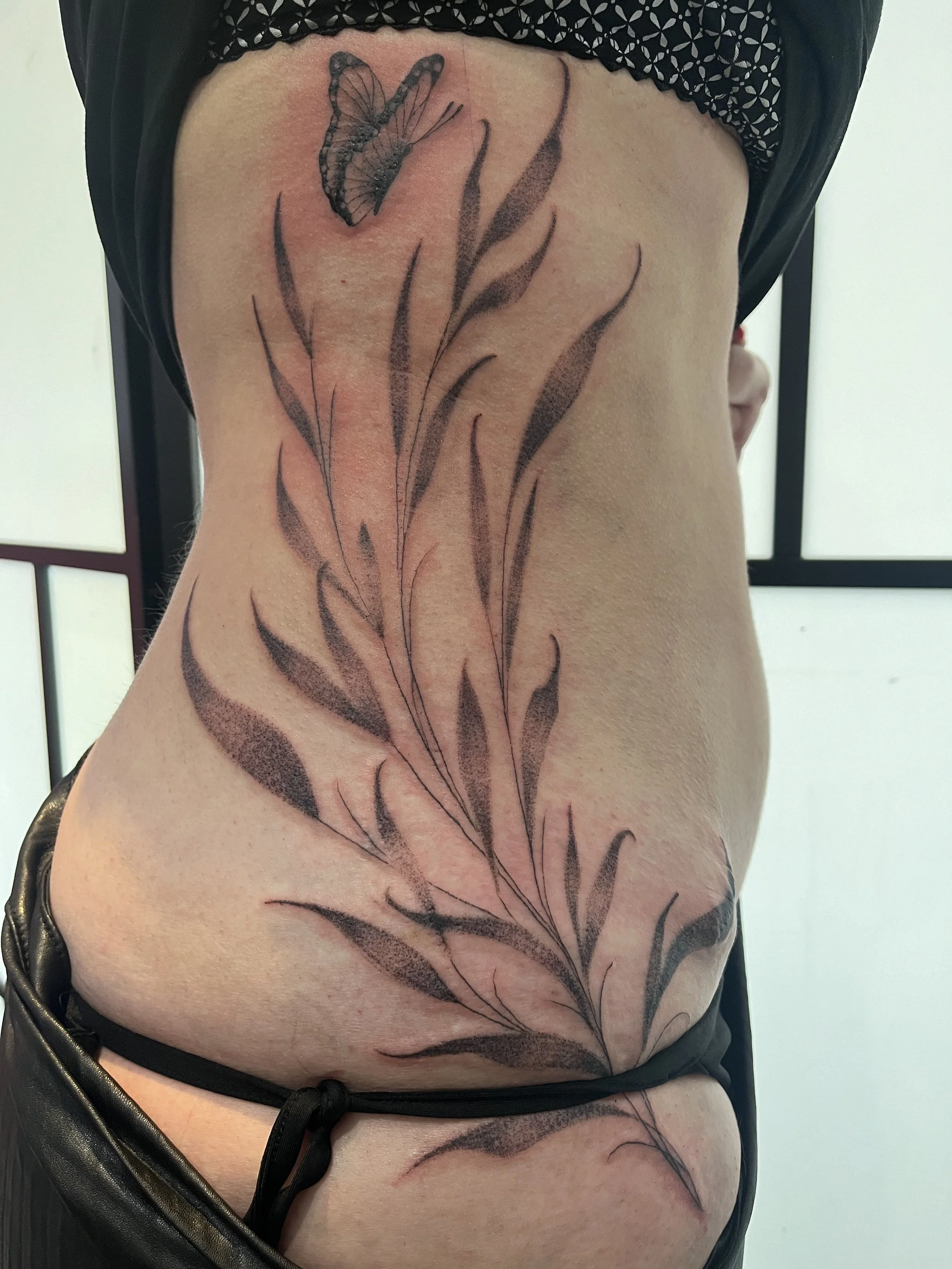 Tatouage floral fine line avec feuillage délicat et papillon réalisé sur les côtes.