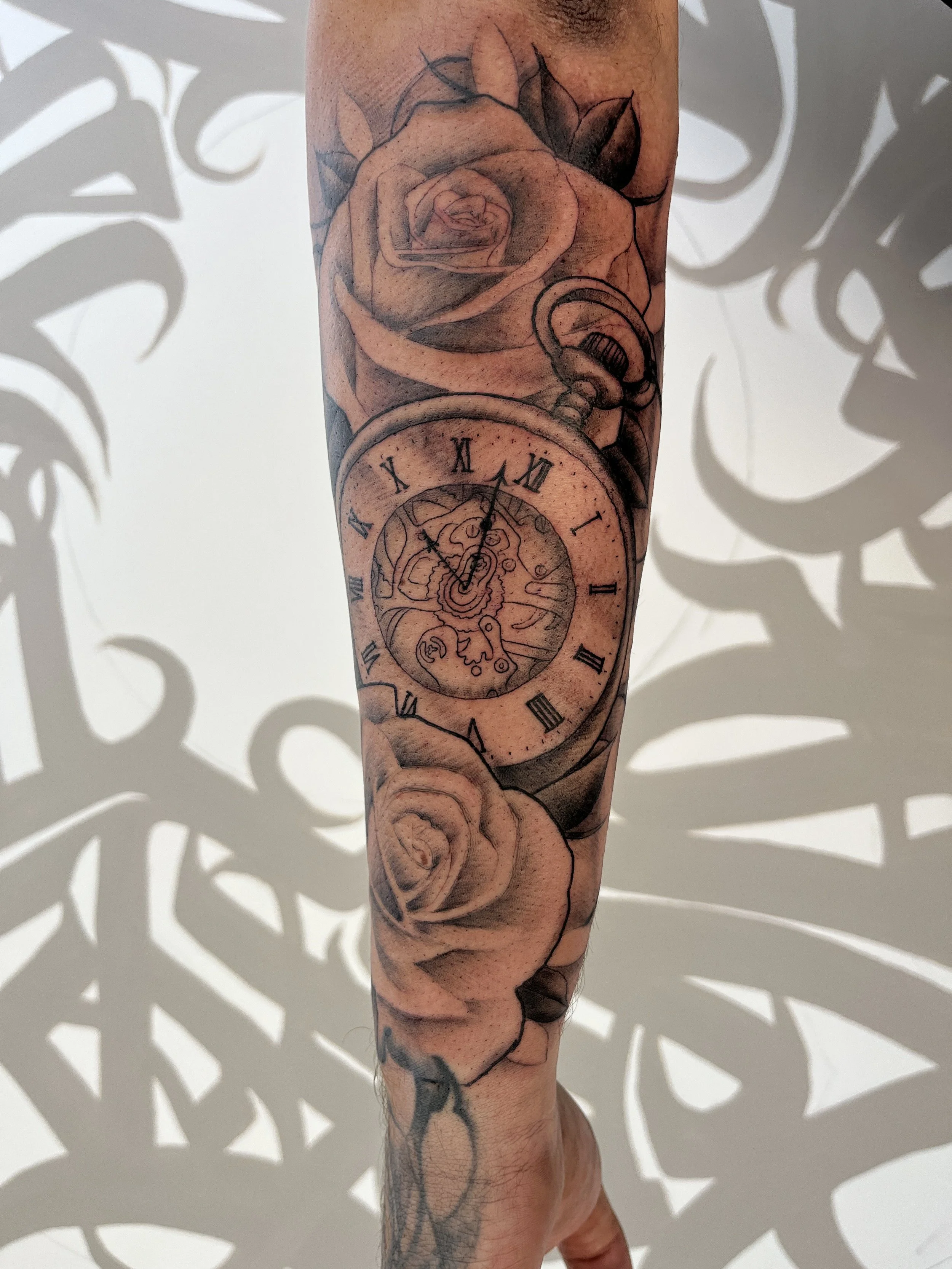 Tatouage réaliste représentant une horloge à mécanisme entourée de roses en noir et gris sur le bras.