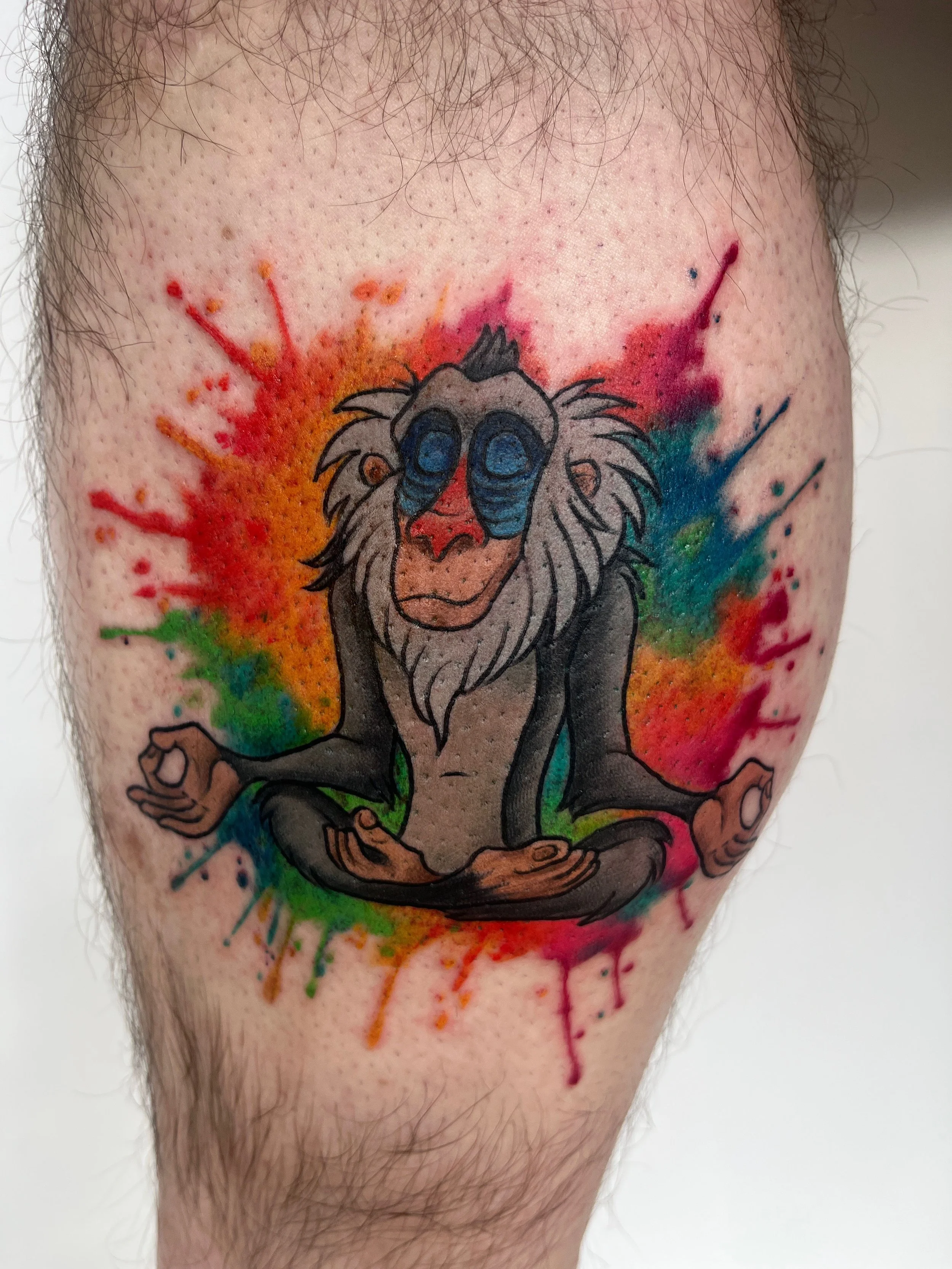 Tatouage cartoon coloré représentant Rafiki du Roi Lion en méditation sur la jambe.