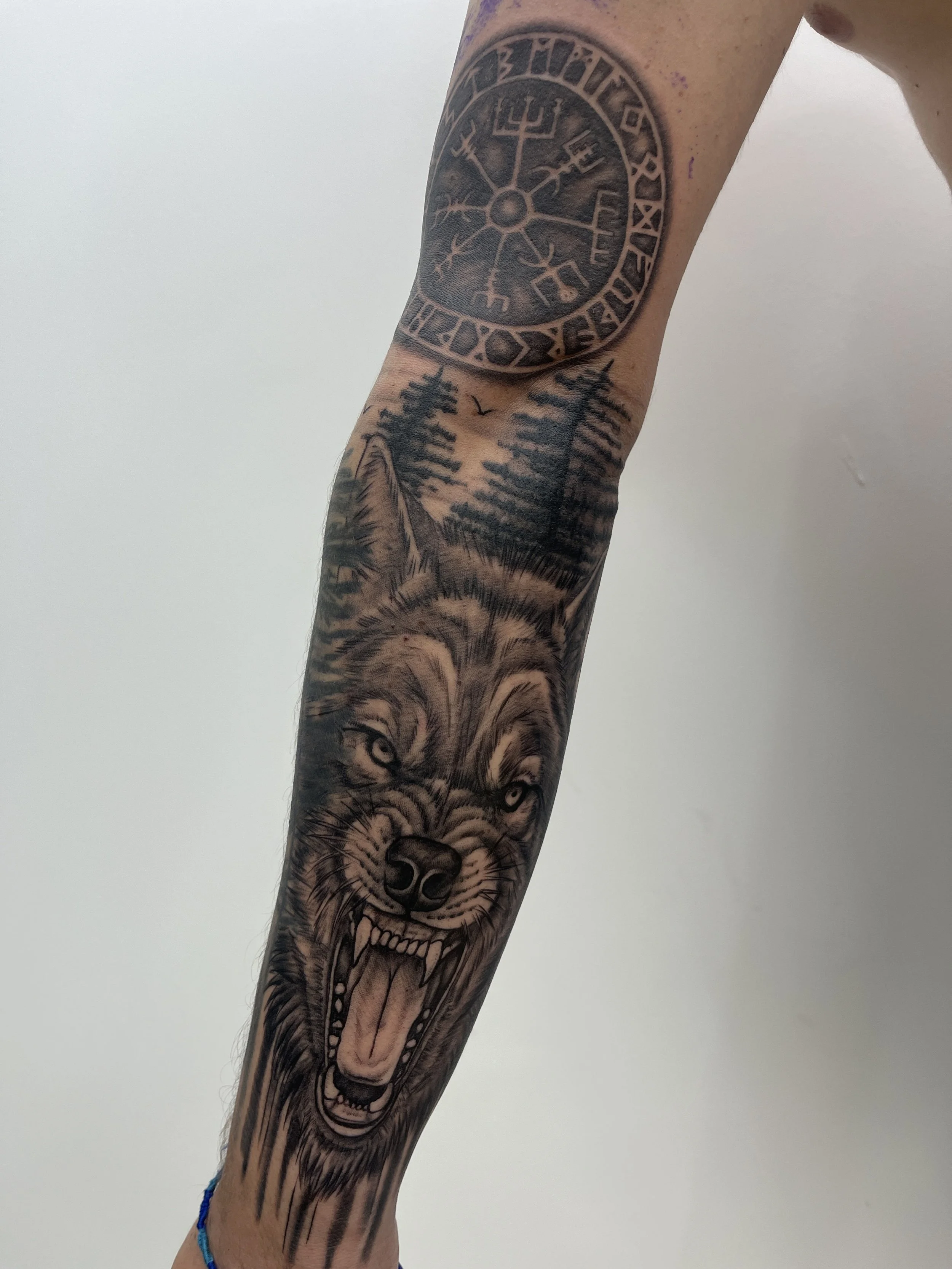 Tatouage style viking représentant un loup hurlant avec forêt nordique et runes sur le bras.