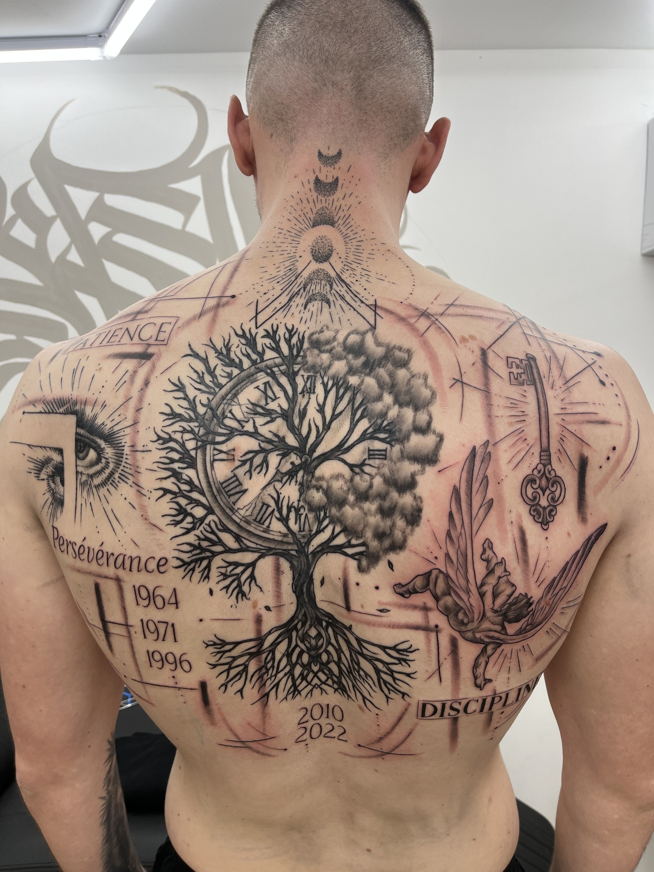 Tatouage graphique et symbolique dans le dos avec arbre, horloge, clé et motifs spirituels.