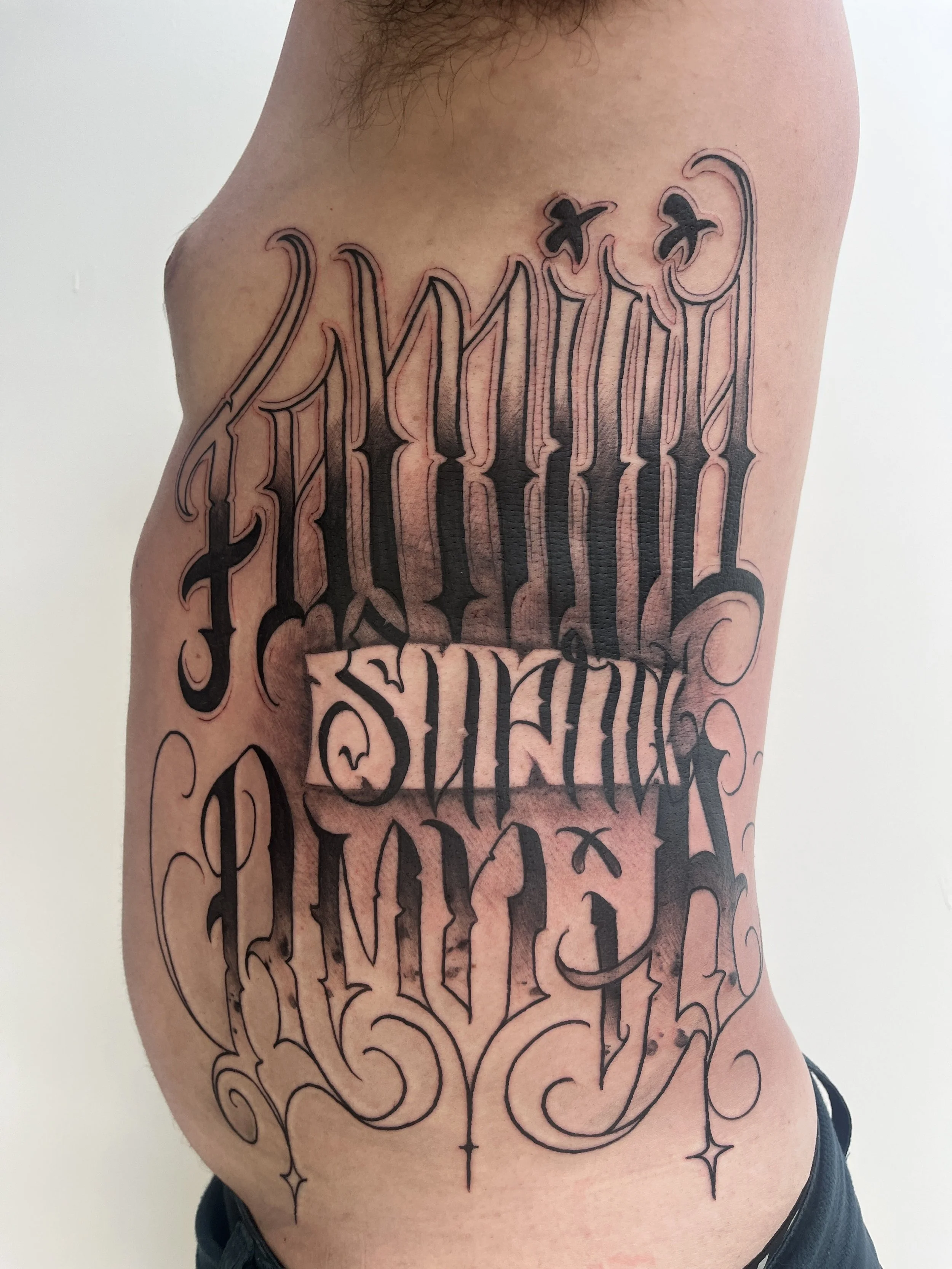 Tatouage lettrage style chicano réalisé sur les côtes avec typographie gothique et ombrages blackwork.