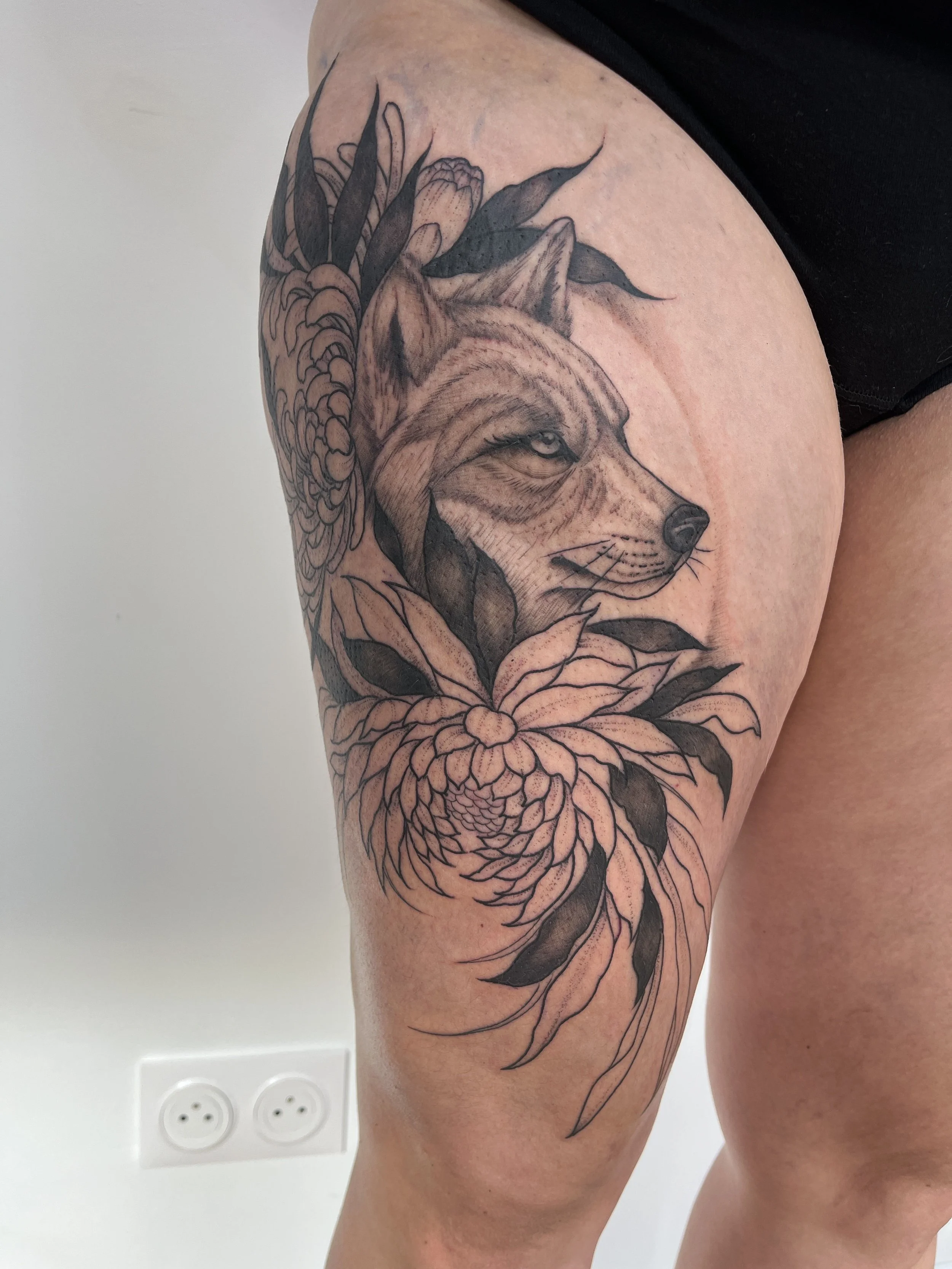Tatouage blackwork représentant un loup entouré de fleurs sur la jambe.