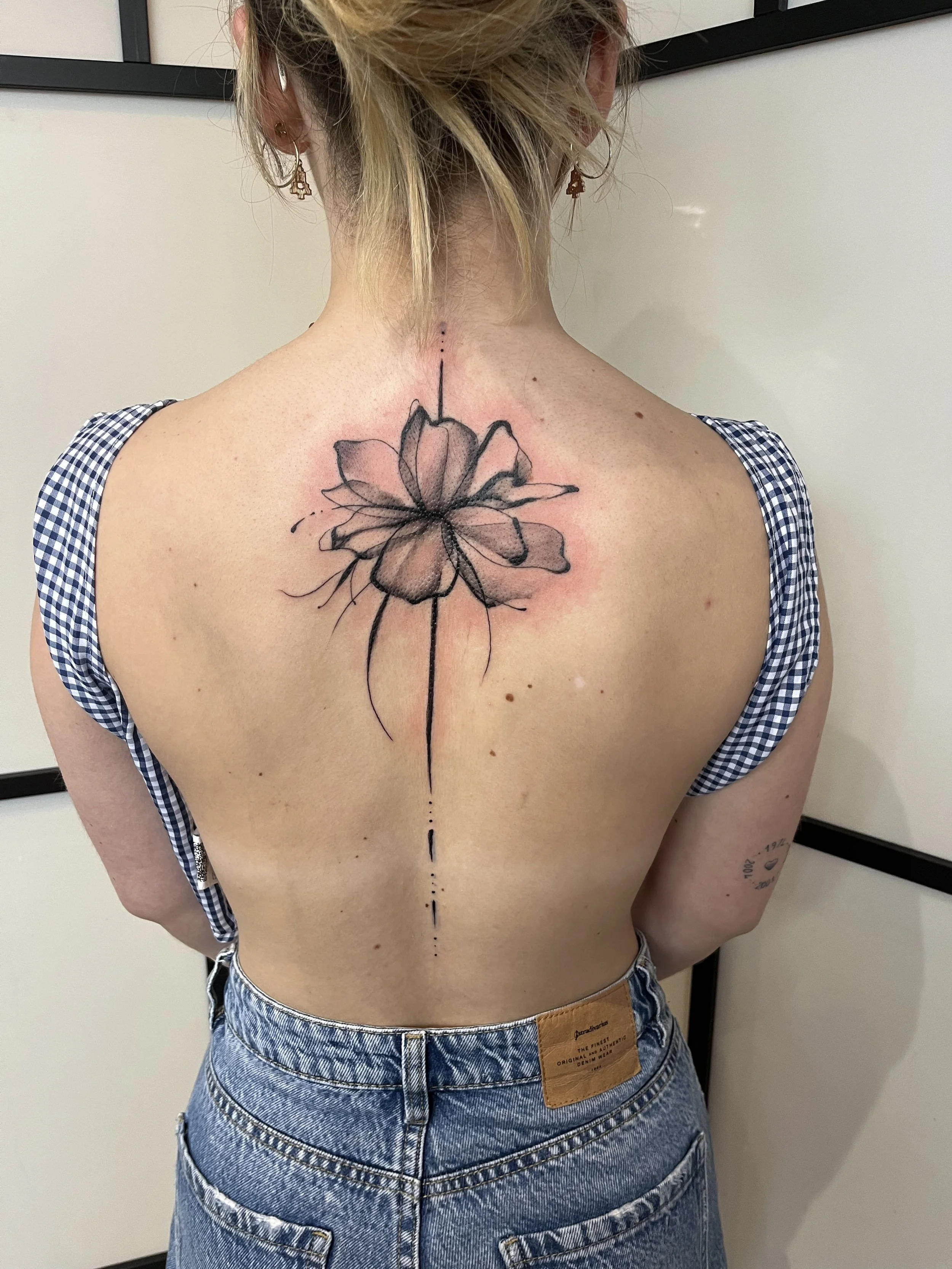 Tatouage graphique floral dans le dos avec fleur centrale et ligne fine verticale minimaliste.