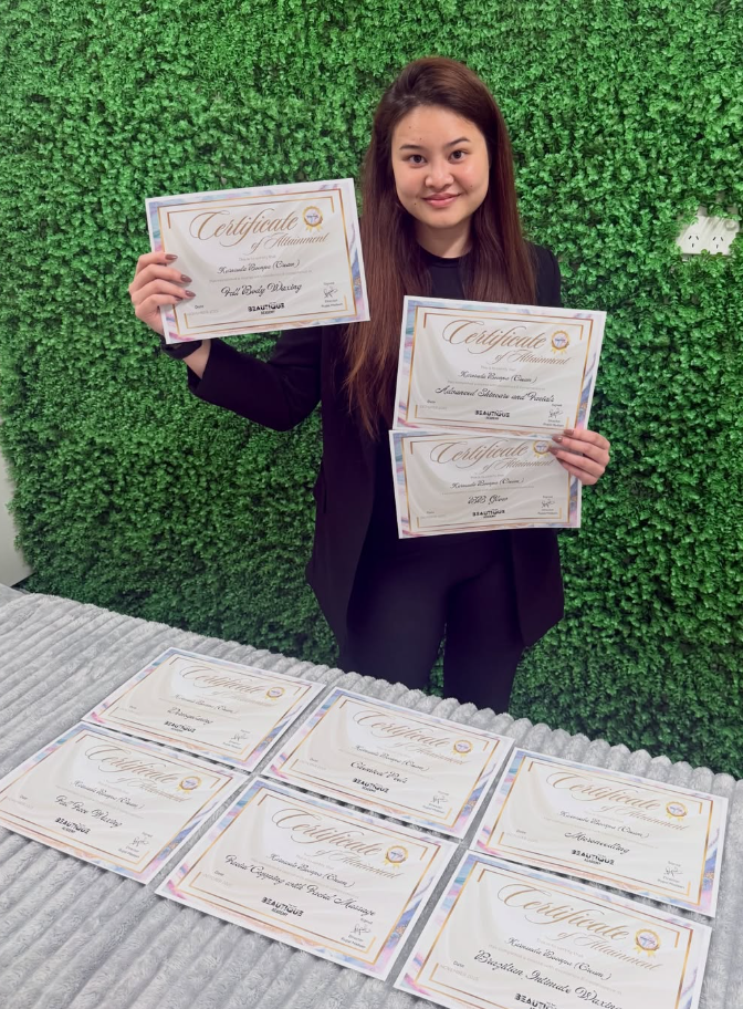 A young woman in a black outfit smiling and holding three certificates of achievement, standing in front of a green hedge wall and a table with multiple certificates laid out.