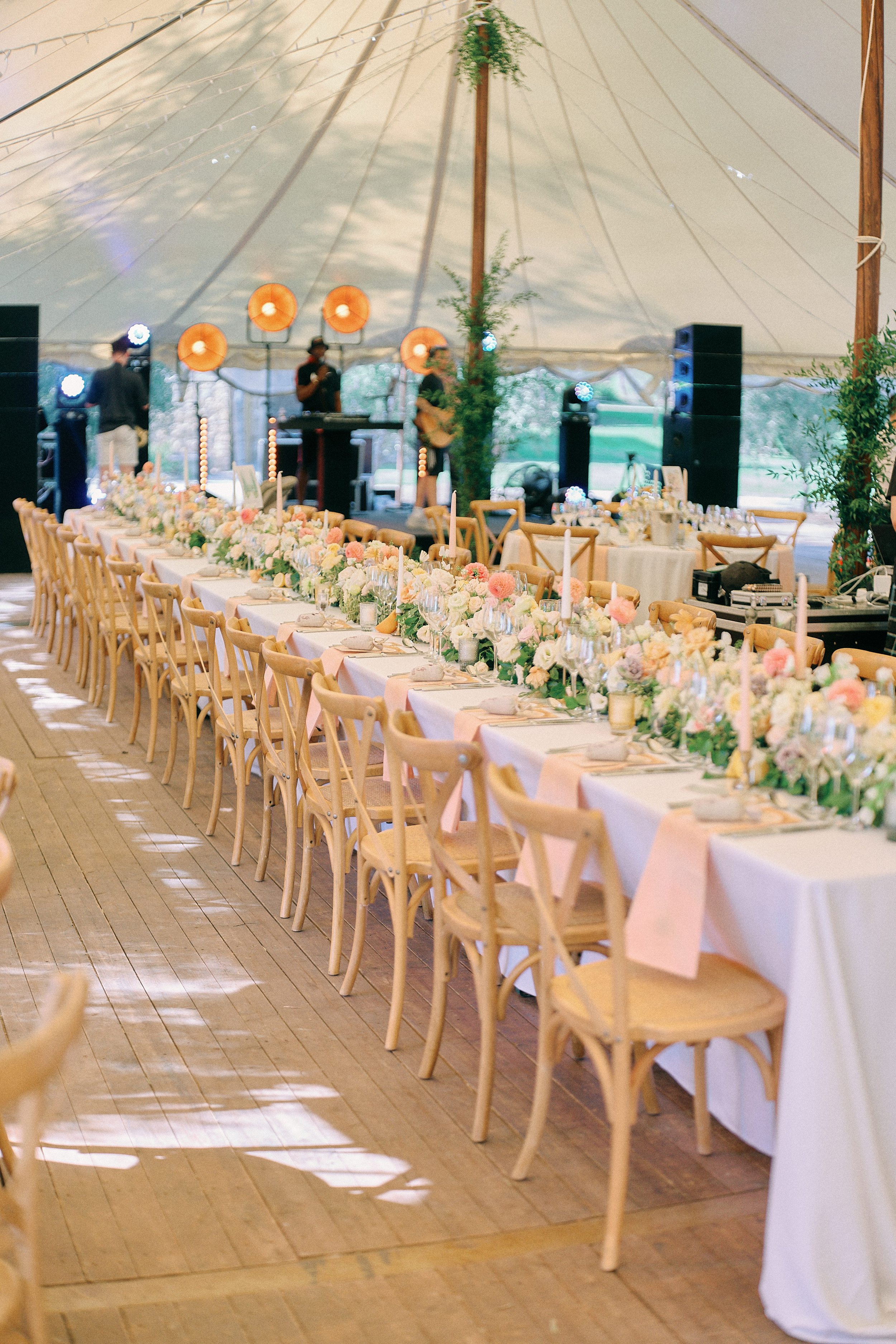 A long banquet table decorated with flowers, candles, and tableware inside a large tent with a live band performing in the background.