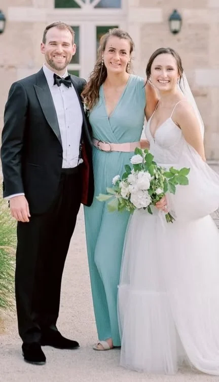 Three people, a man in a tuxedo, and two women, one in a light blue dress and the other in a wedding gown, standing outside a building with a bouquet of flowers.