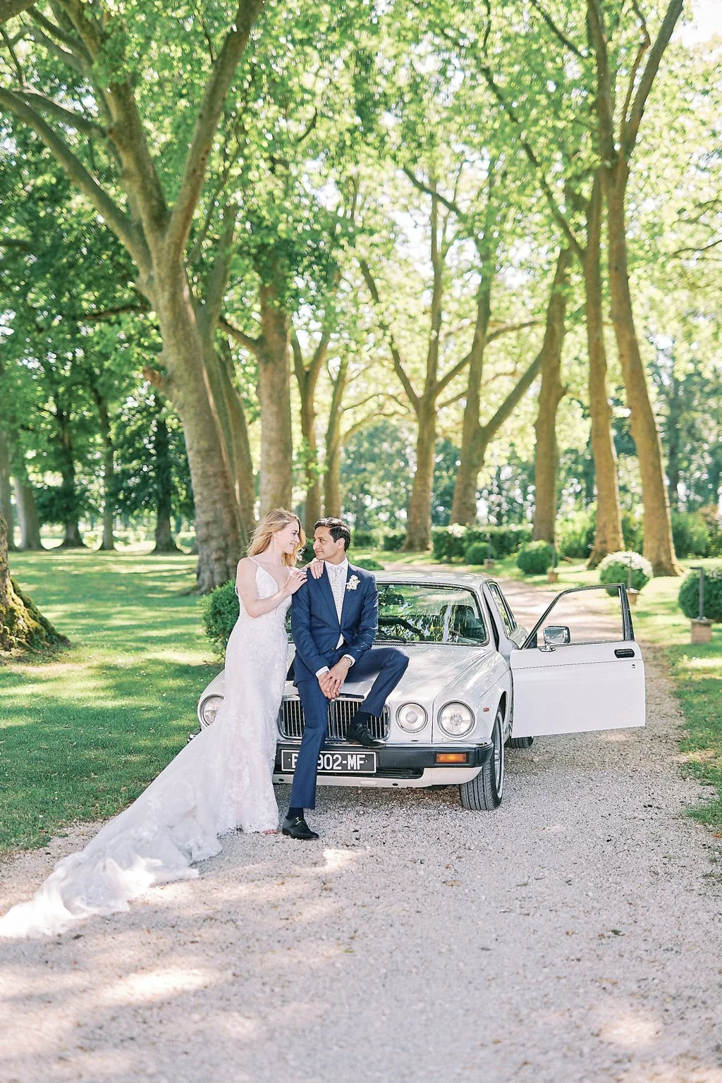 A bride in a white wedding dress and a groom in a blue suit with a white shirt and boutonniere sitting on the hood of a white vintage car in a park with tall trees and sunlight filtering through the leaves.