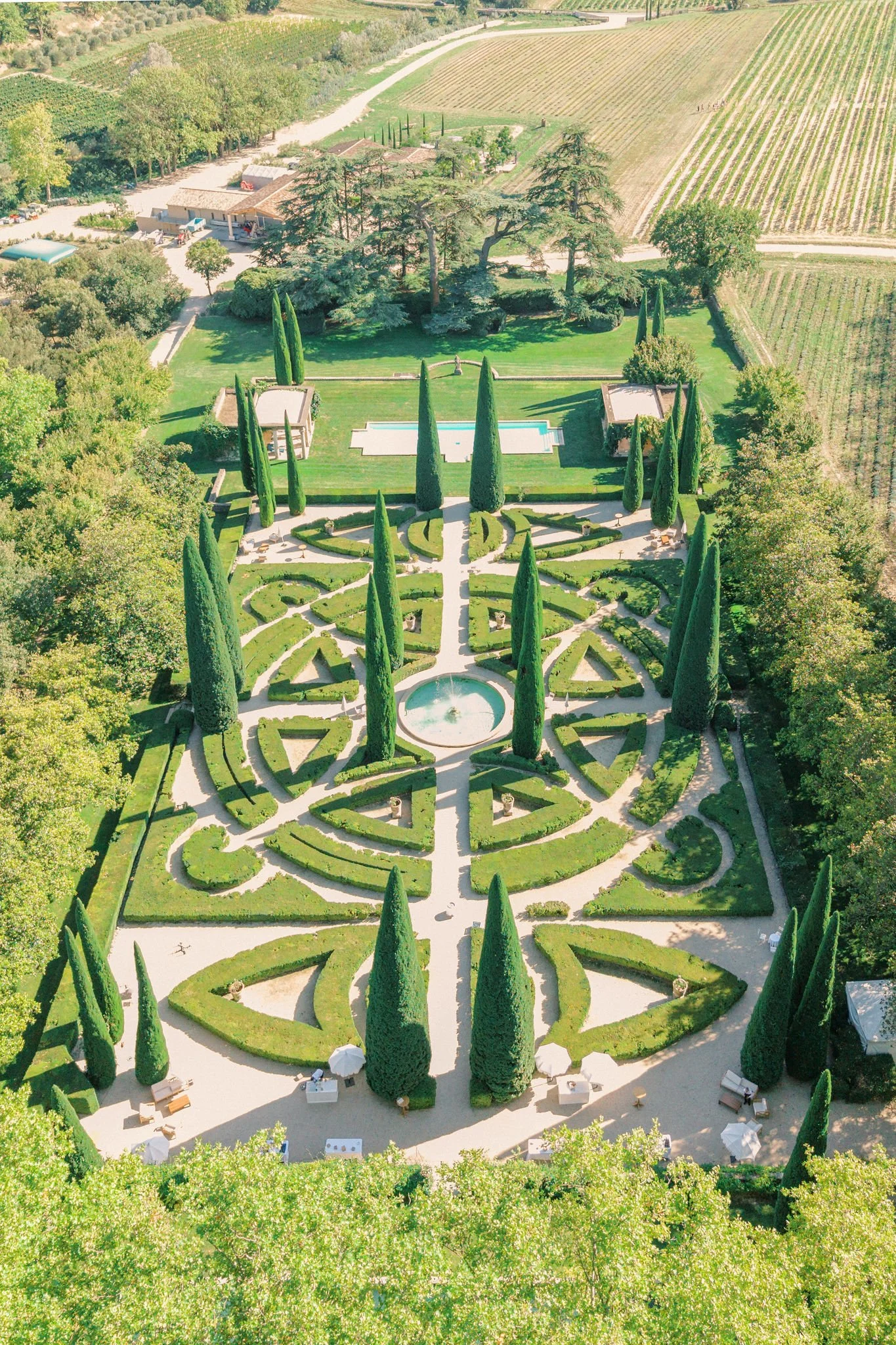 Aerial view of a formal garden with symmetrical layout, tall cypress trees, a fountain at the center, and surrounding pathways and seating areas, with vineyards and rural landscape in the background.