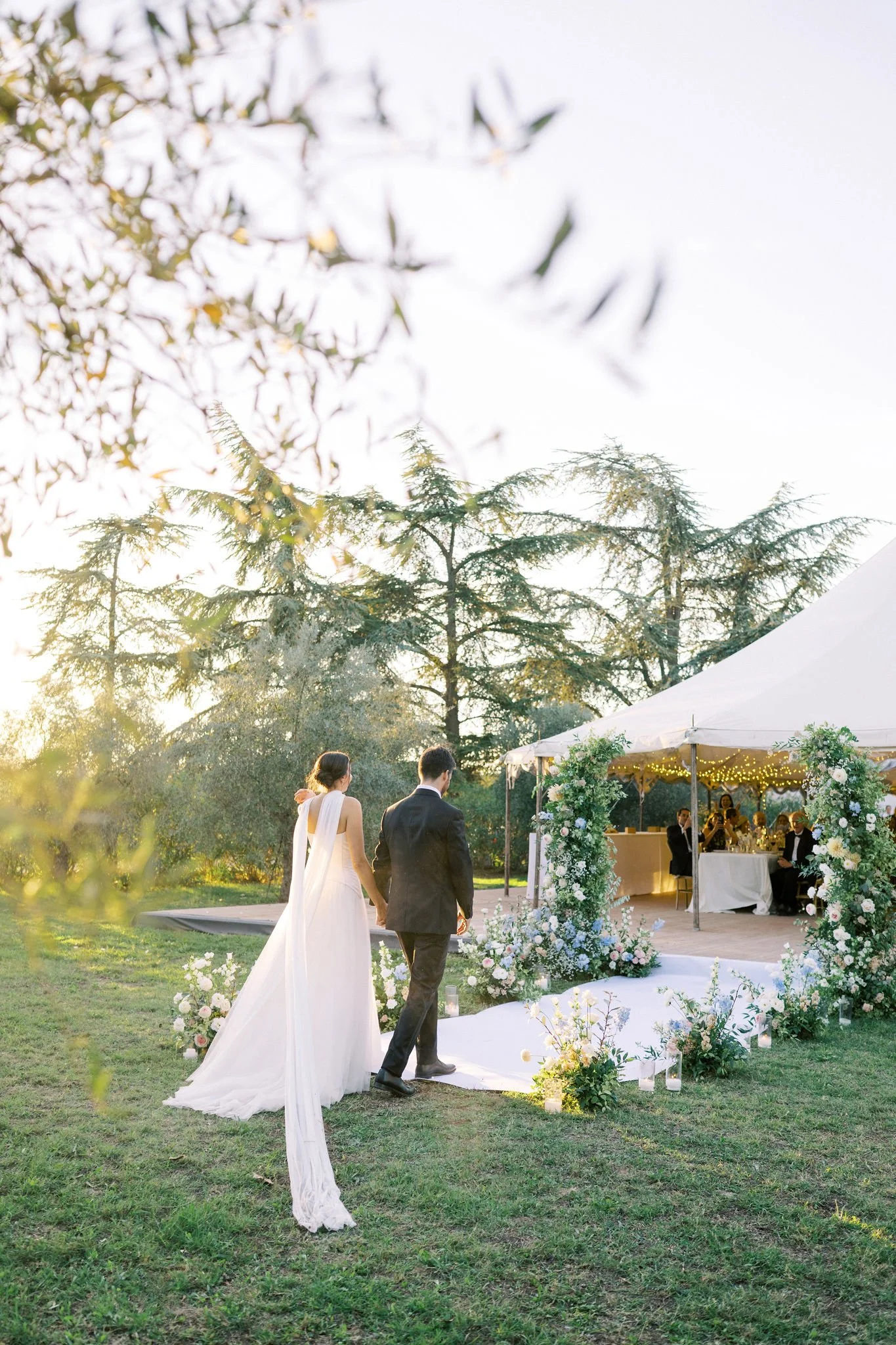 Bride and groom walking hand in hand towards a decorated outdoor wedding altar during sunset in a garden setting.