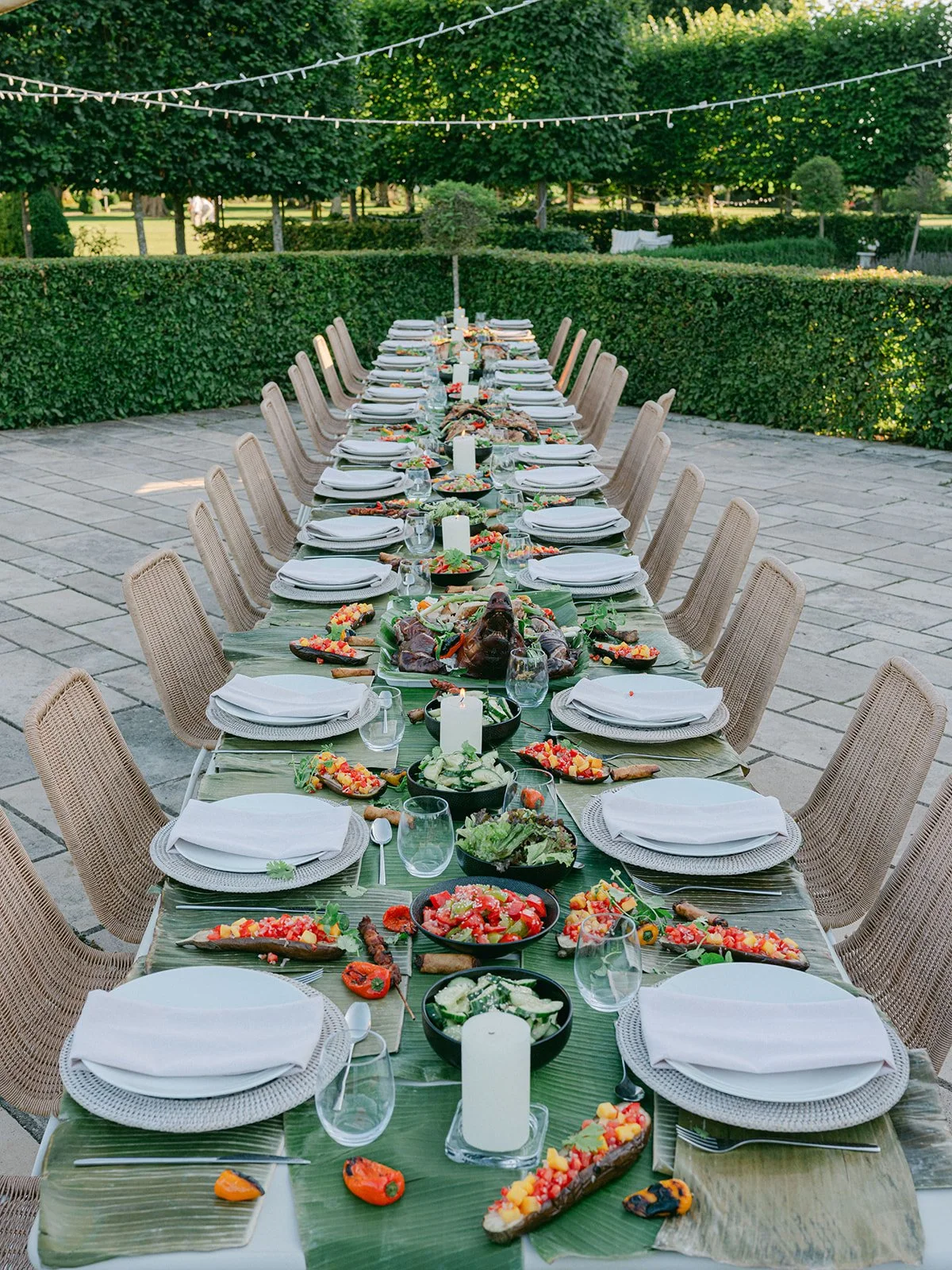 Long outdoor dining table set for a meal with plates, glasses, napkins, and food, decorated with candles and surrounded by wicker chairs on a stone patio with greenery and string lights overhead.