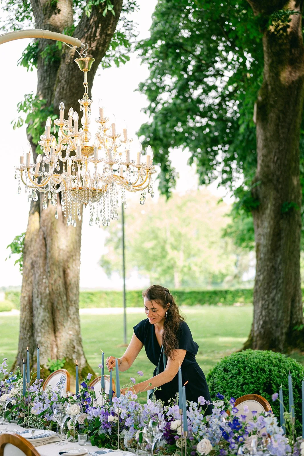 Woman decorating a table with purple and white flowers outdoors, hanging a blue candle, with large trees and a chandelier overhead.