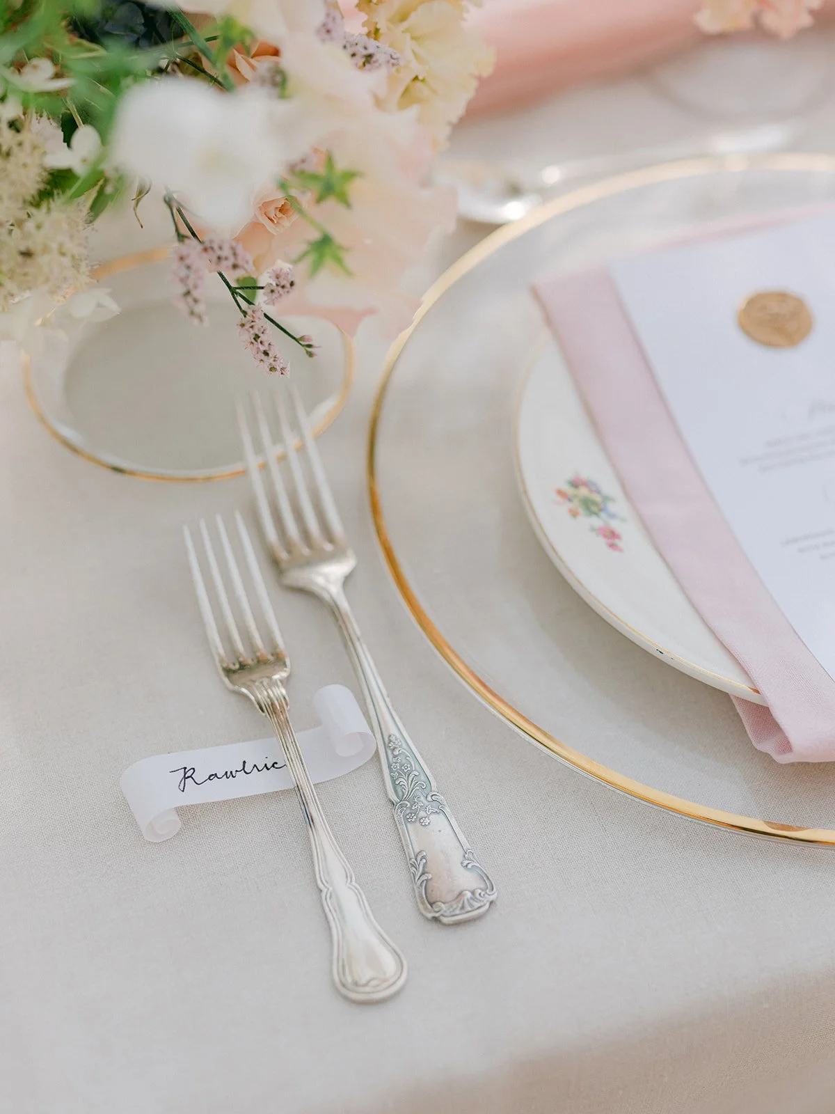 Elegant table setting with two silver forks, a floral centerpiece, a pink and gold-rimmed plate, and a menu card with a pink napkin.