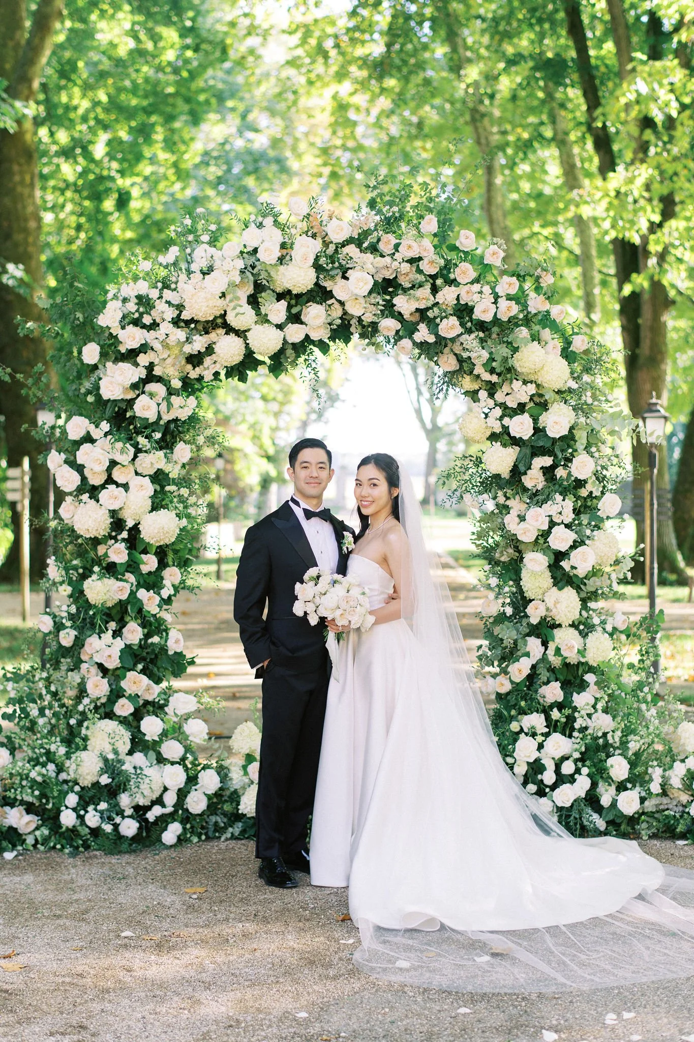 A bride and groom standing under a floral arch in a park, smiling for a wedding photo.