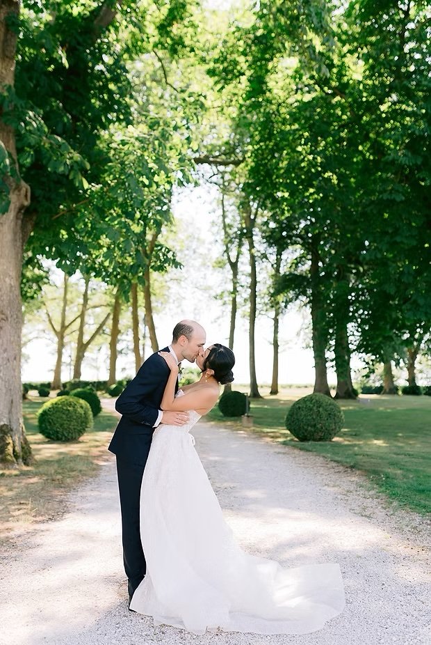 A newlywed couple sharing a kiss outdoors on a tree-lined path with lush green foliage.