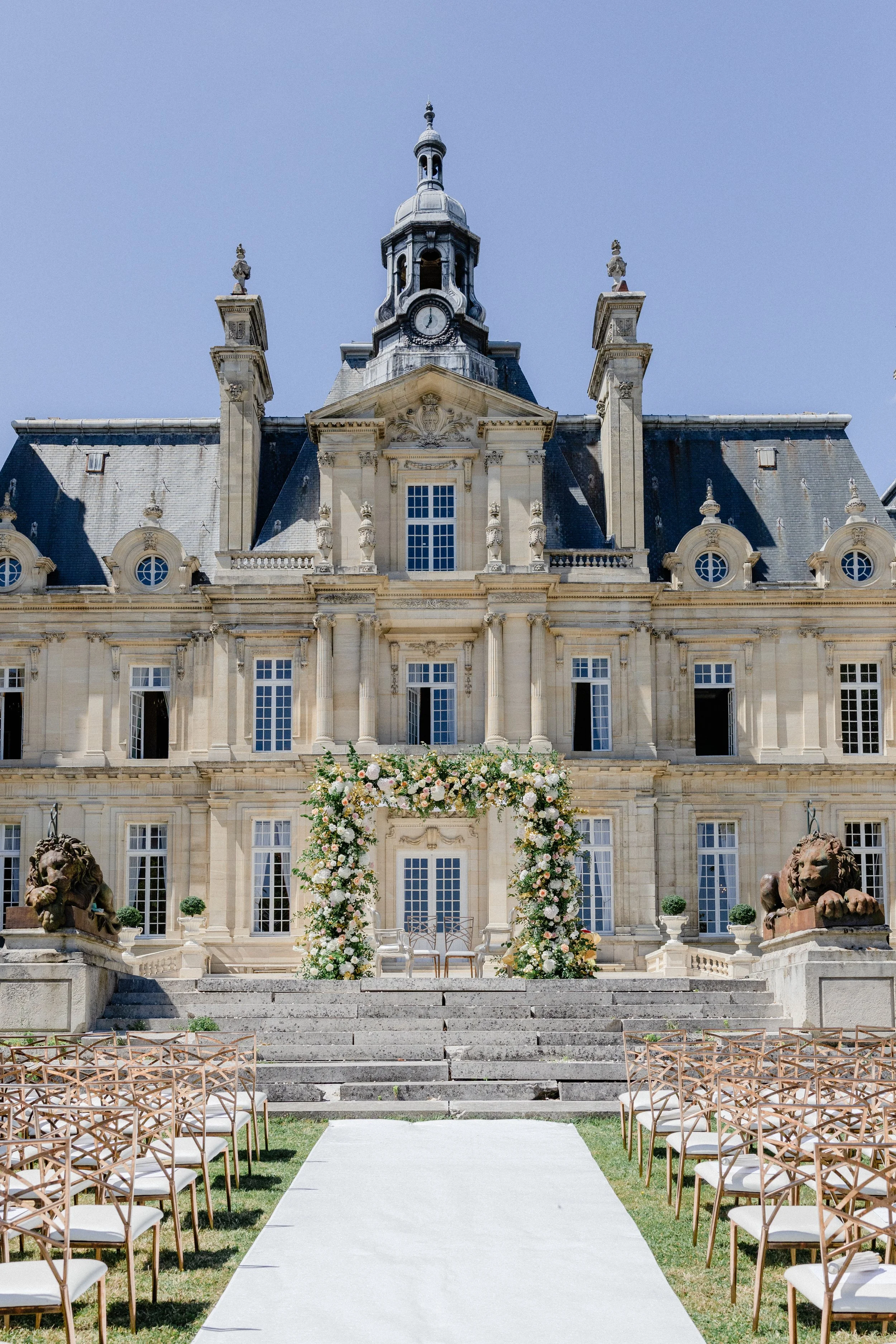 Outdoor wedding setup in front of a historic mansion with a floral arch, chairs, and lion statues.