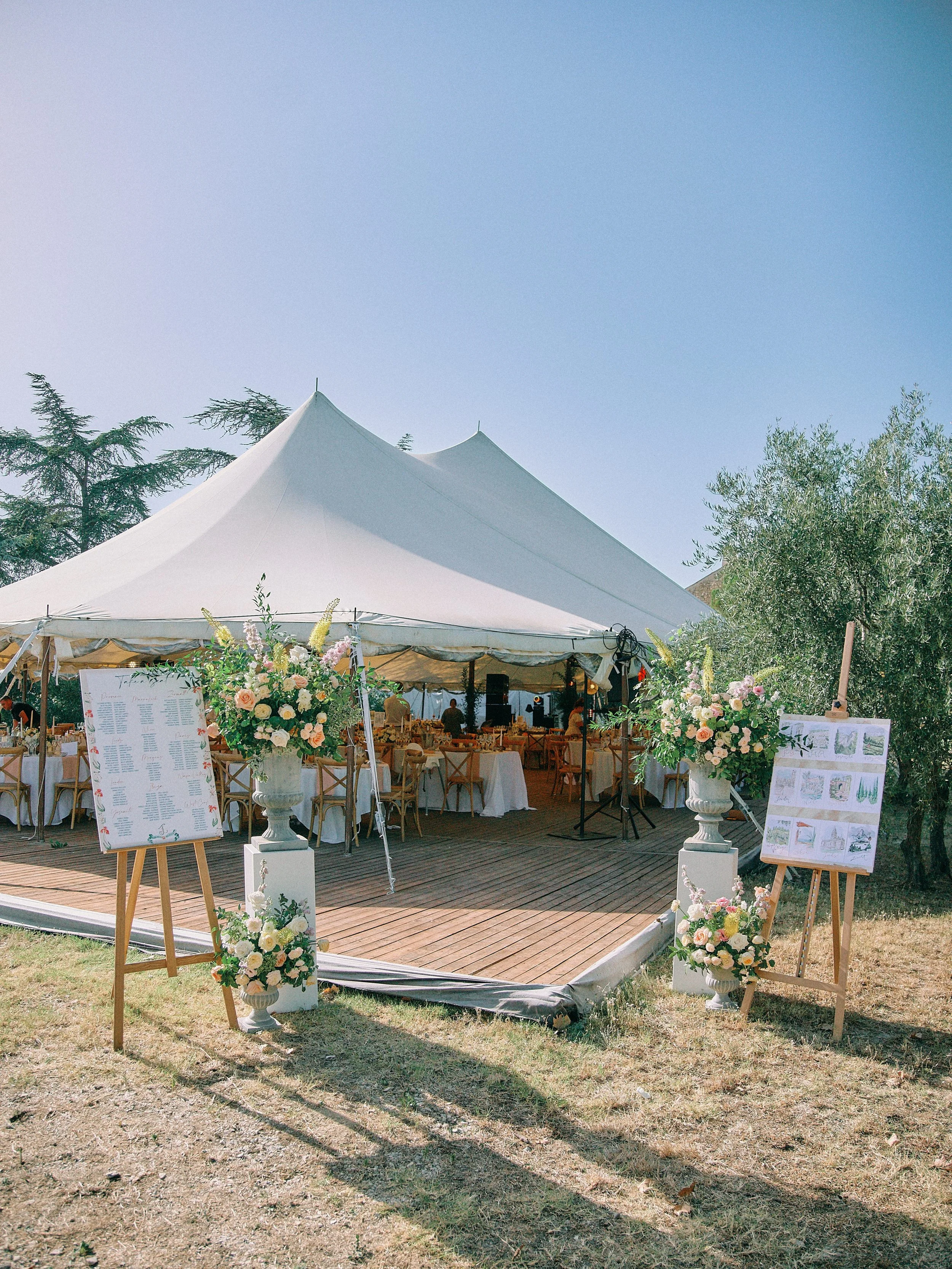 Outdoor wedding reception under large white tent with floral arrangements on pedestals and seating for guests.