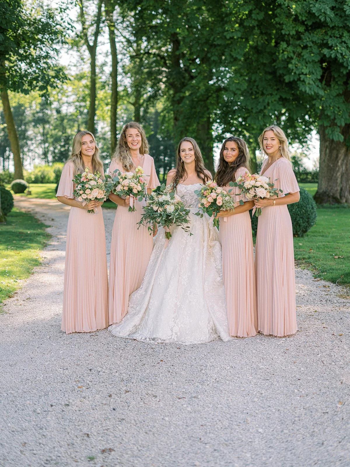A bride and four bridesmaids standing outdoors on a gravel path, holding bouquets of flowers, with green trees and grass in the background.