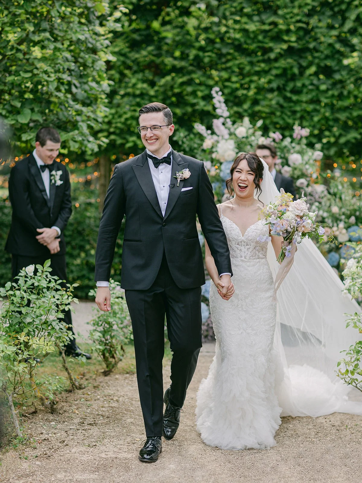 A smiling bride and groom walking hand in hand outdoors at their wedding, with guests and floral decorations in the background.
