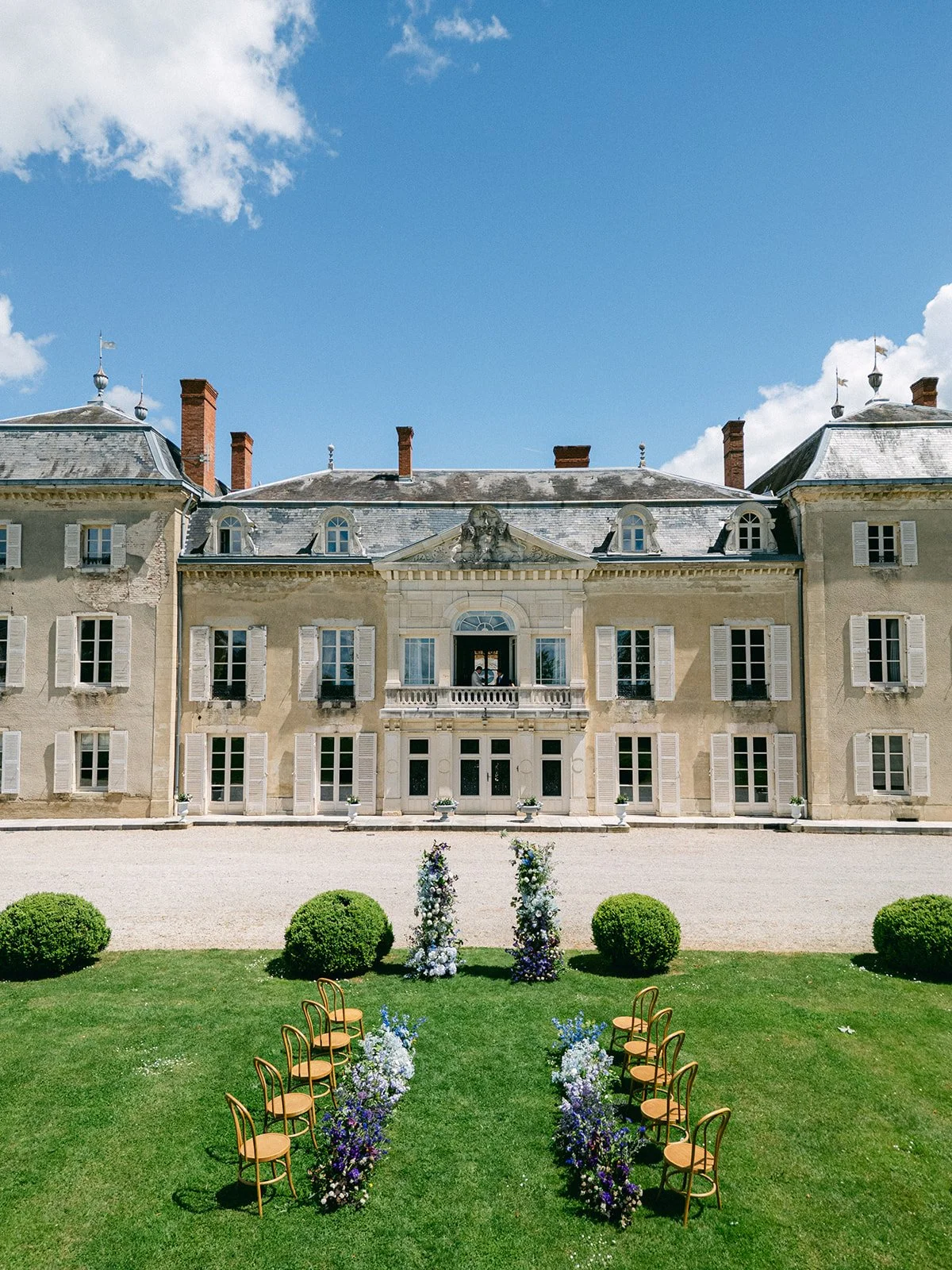An elegant outdoor wedding ceremony setup in front of a historic mansion, with chairs arranged on a grassy lawn and floral decorations along the aisle.