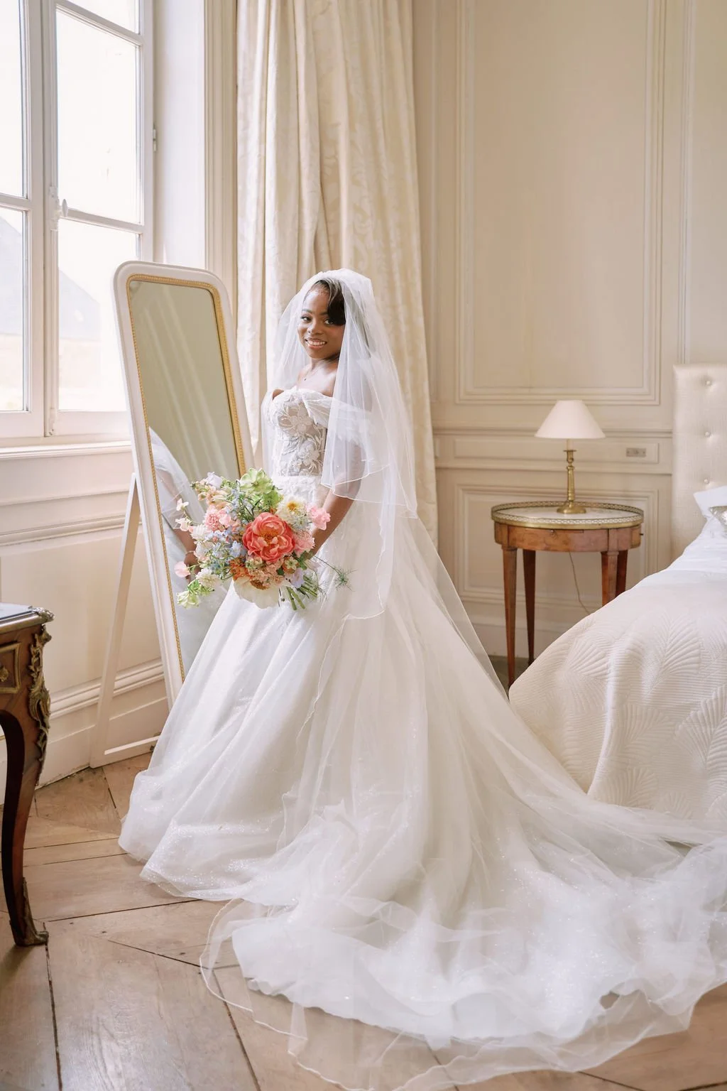 A bride in a white wedding dress with a veil, holding a bouquet of pink and white flowers, standing in front of a mirror in a well-lit, elegant bedroom with cream-colored walls, a gold lamp on a side table, and a bed with white linens.