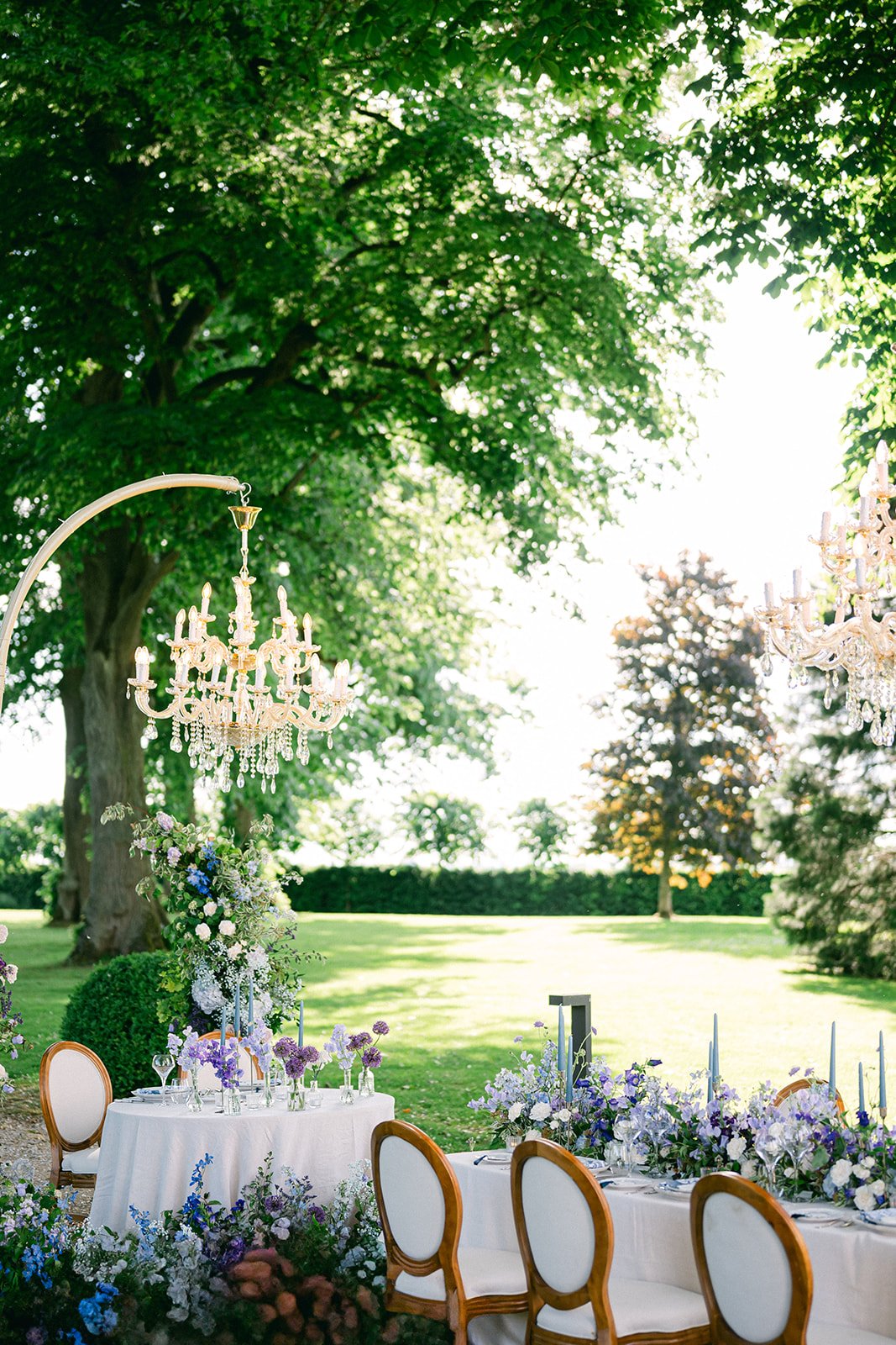 An outdoor event setup with a long table decorated with purple and white flowers, surrounded by chairs with white upholstery and wooden frames. There is a smaller round table with similar decorations. Elegant chandeliers hang from above, set against 