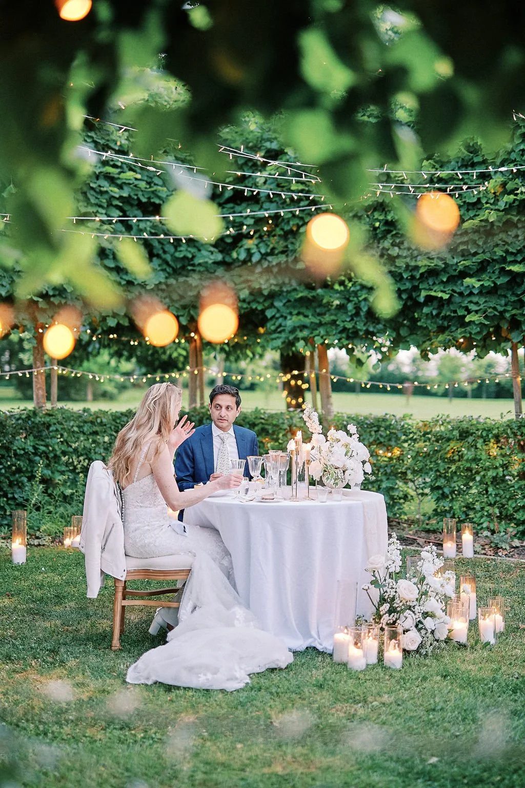A bride and groom having a conversation at an outdoor wedding reception table under trees decorated with string lights and lanterns, with candles and white flowers on the table and around on the grass.