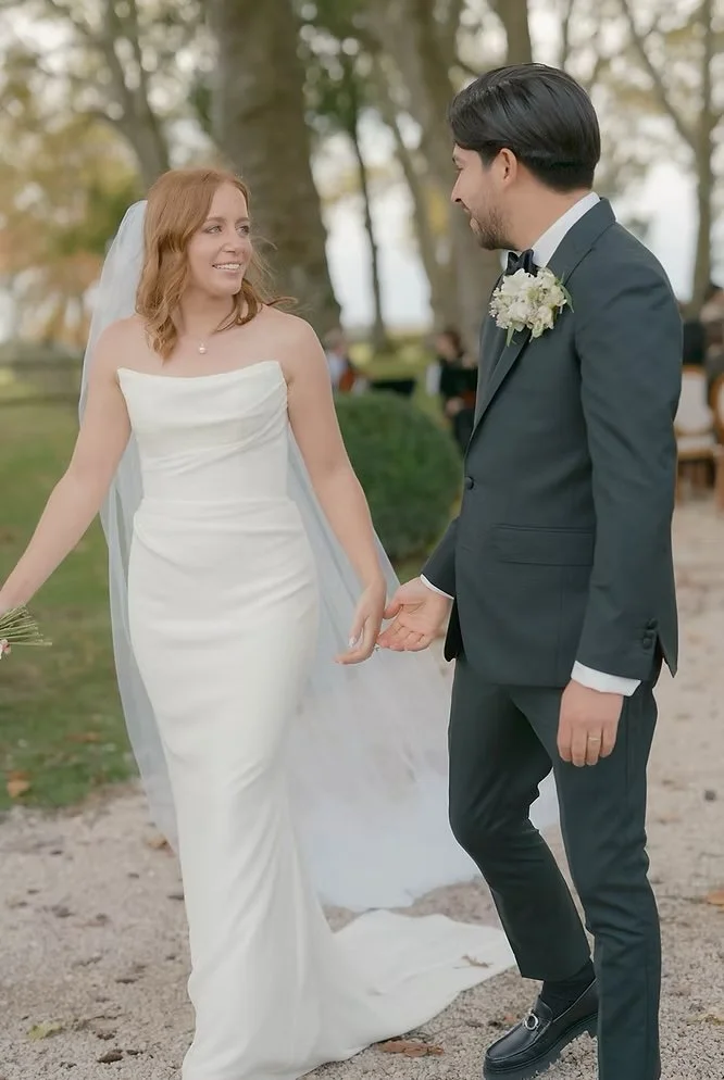 A bride and groom holding hands and smiling at each other outdoors during a wedding ceremony.