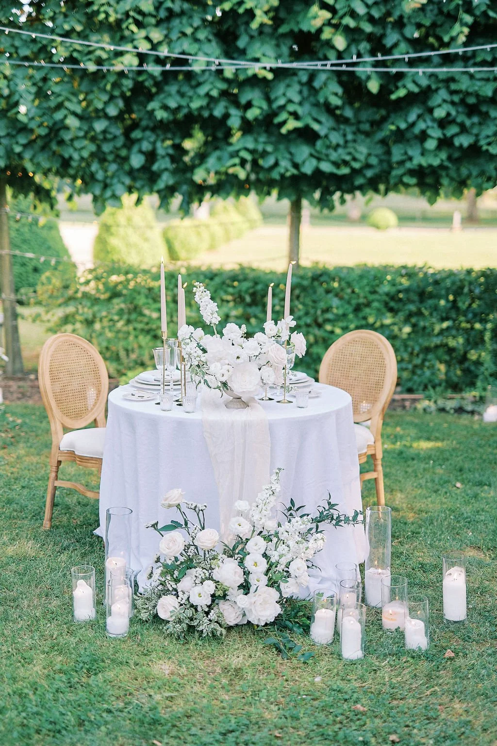 Elegant outdoor wedding table decorated with white flowers, candles, and tall candlesticks, set in a lush garden.