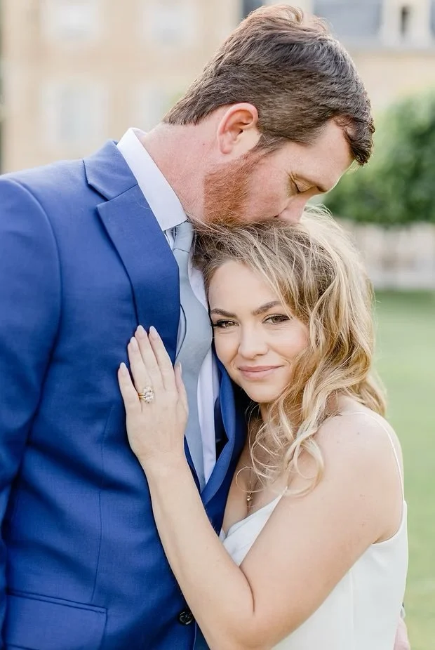 A man in a blue suit and a woman with long blonde hair are embracing outdoors, with the man gently resting his head on the woman's head. The woman is smiling softly, and they look happy and close.