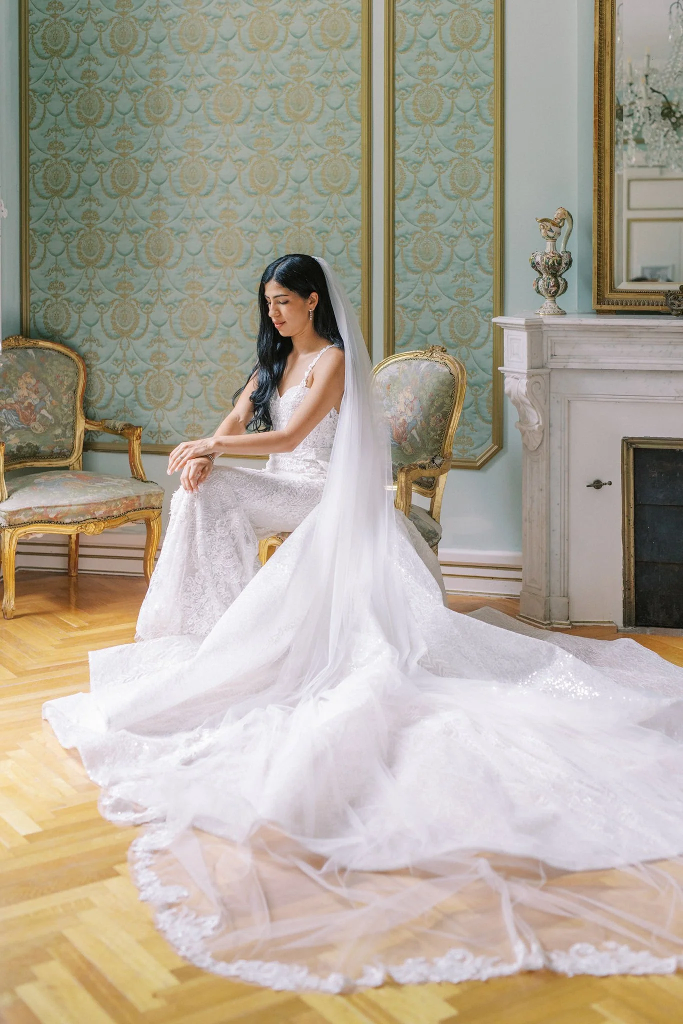 A bride sits on a vintage-style armchair in an elegant room, wearing a white lace wedding gown with a long train and a veil, with a marble fireplace and ornate wallpaper in the background.