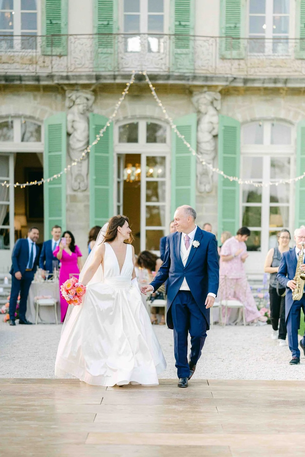 A bride in a white wedding gown holding a pink flower bouquet, walking with a groom in a dark blue suit at an outdoor wedding reception, with guests in colorful dresses and suits in the background, and a grand building with green shutters behind them