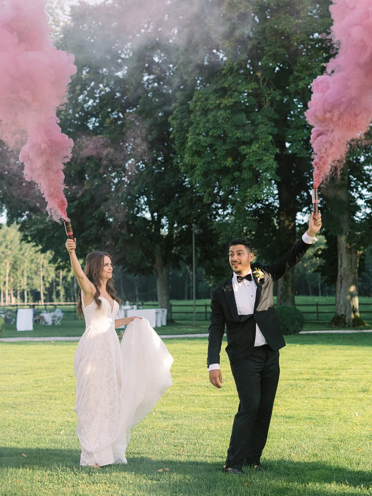 Bride and groom in wedding attire holding pink smoke bombs outdoors on green grass, with trees and tables in the background.