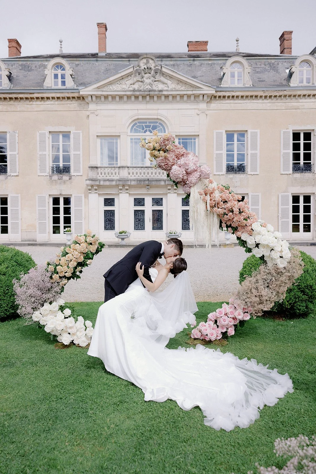 A bride and groom sharing a kiss in front of a floral arch on a lawn in front of a large, ornate building.