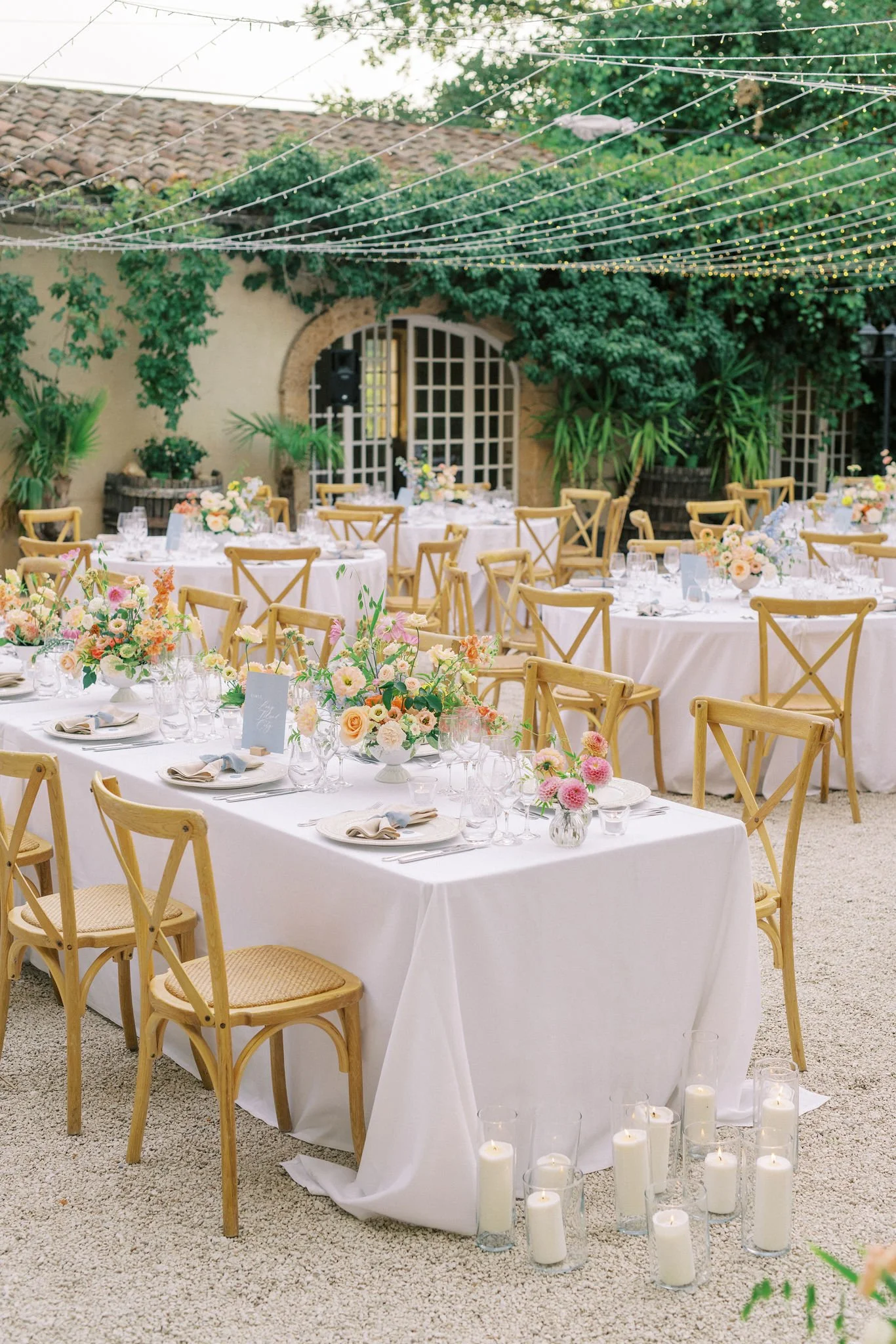 Elegant outdoor wedding reception with round and rectangular tables covered in white tablecloths, decorated with pastel flower centerpieces, set with glassware and napkins, surrounded by wooden chairs, surrounded by lush green plants and string light