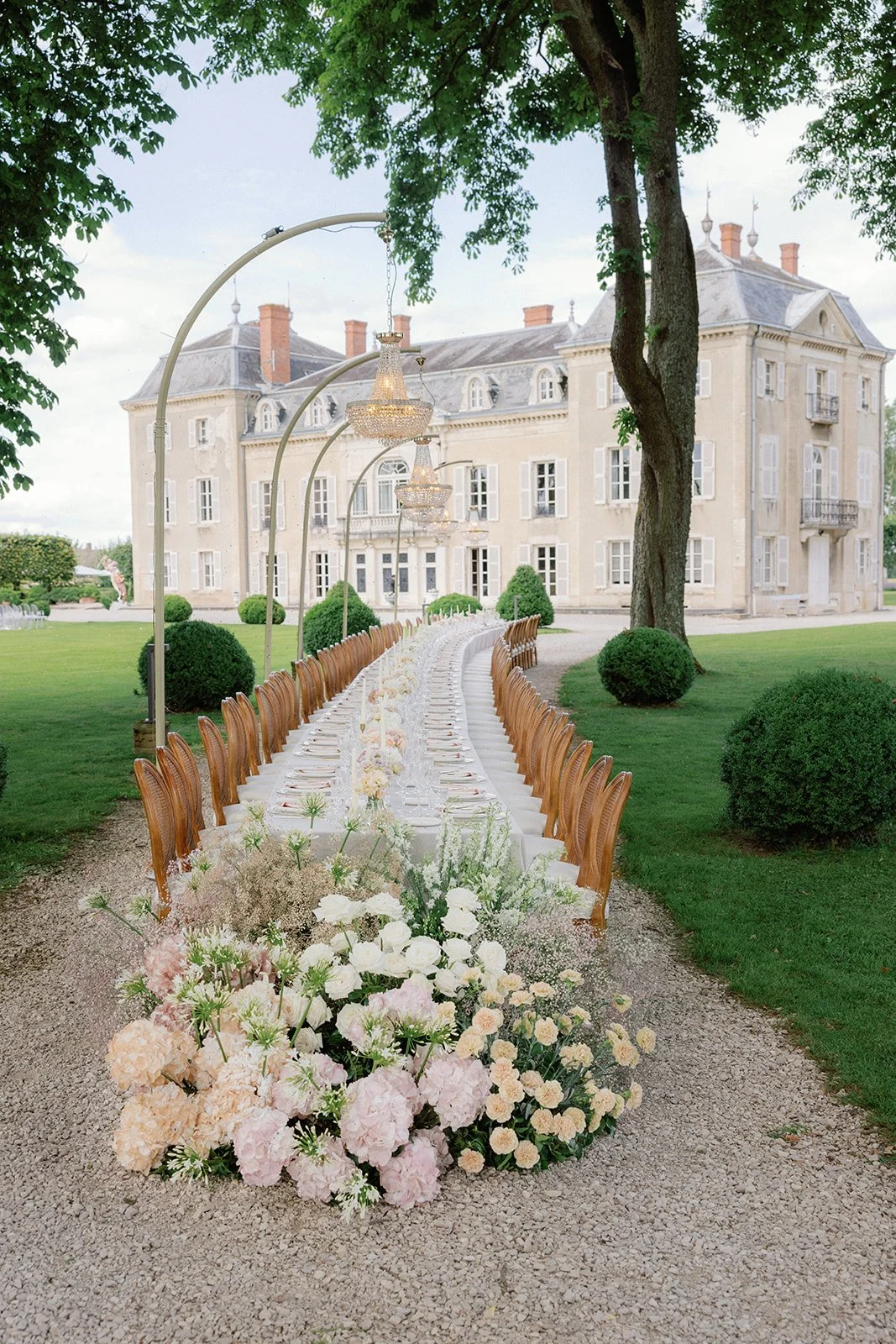An outdoor wedding table set up with floral arrangements, elegant chandeliers, and chairs, in front of a large historic mansion surrounded by green lawns and trees.