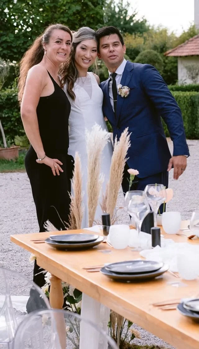 Three people standing close together at an outdoor event, with a table set with plates, glasses, and decorations in the foreground.