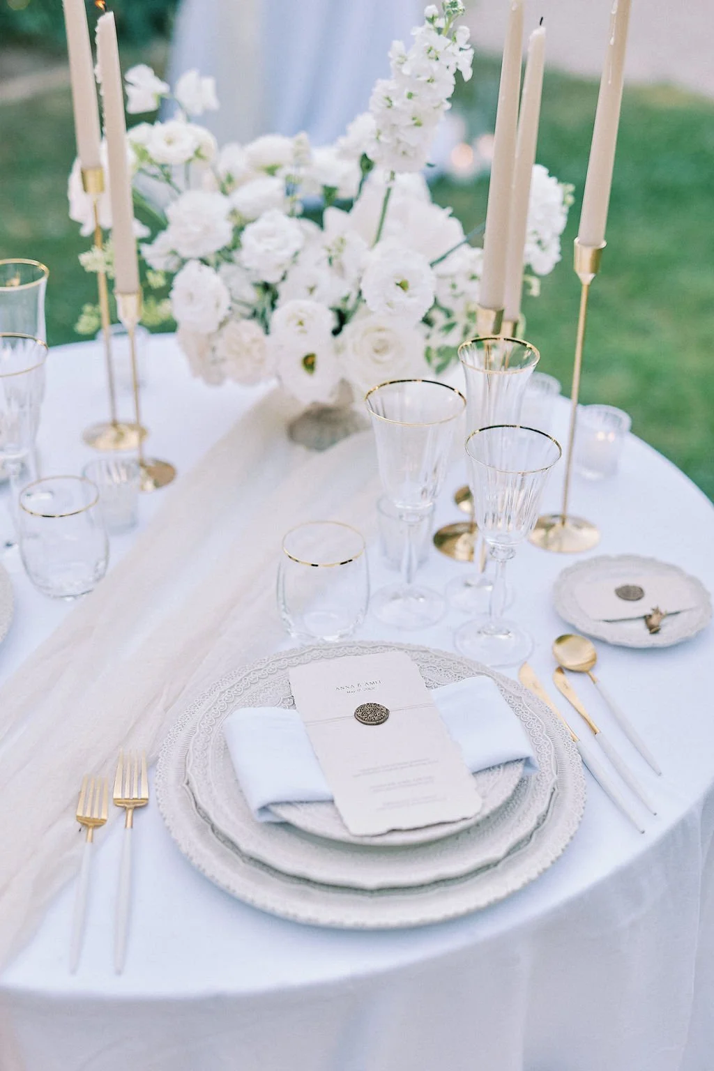 Elegant outdoor table setting with white plates, gold cutlery, glassware with gold rims, a menu with a wax seal, and a centerpiece of white flowers and tall candles.